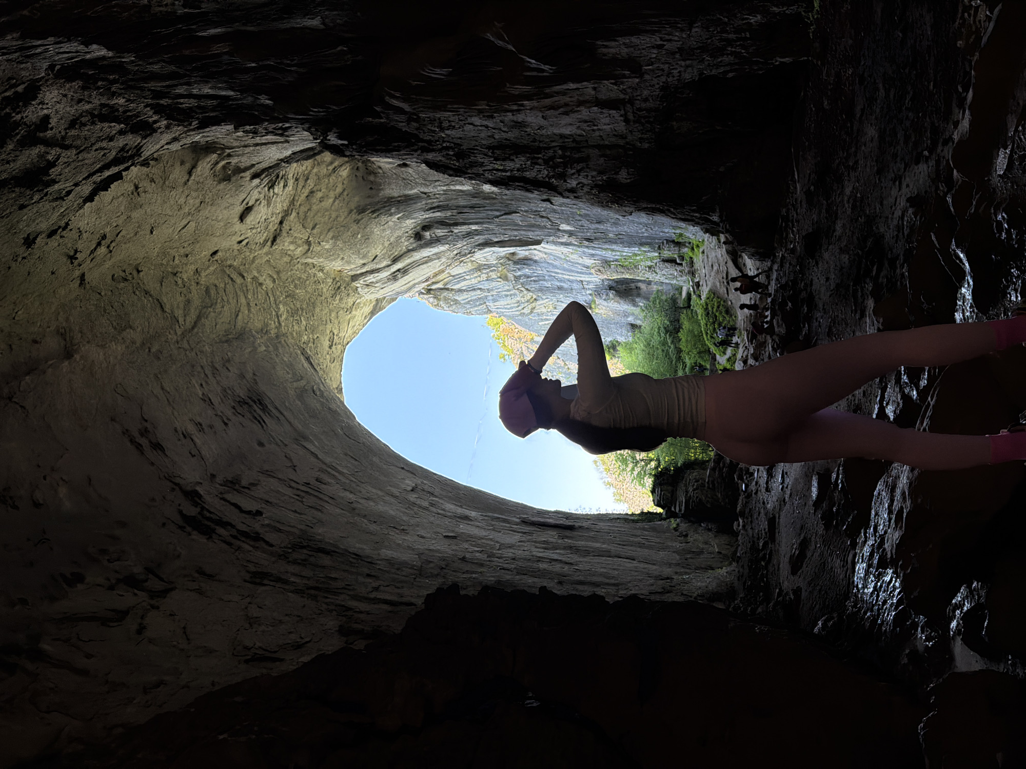 A low-angle shot of a person inside a cave, identified as Devetashka Cave in Karlukovo, Bulgaria. The person is standing near the cave entrance, facing the opening where the sky is visible, suggesting daytime. The cave walls are rocky and partially illuminated, contrasting with darker, shadowed areas. The person is wearing what appears to be a light-colored outfit and a hat, with arms raised towards their head. Outside the cave entrance, some greenery and vegetation are visible. The scene captures the perspective from within the cave looking out towards the natural light and landscape.