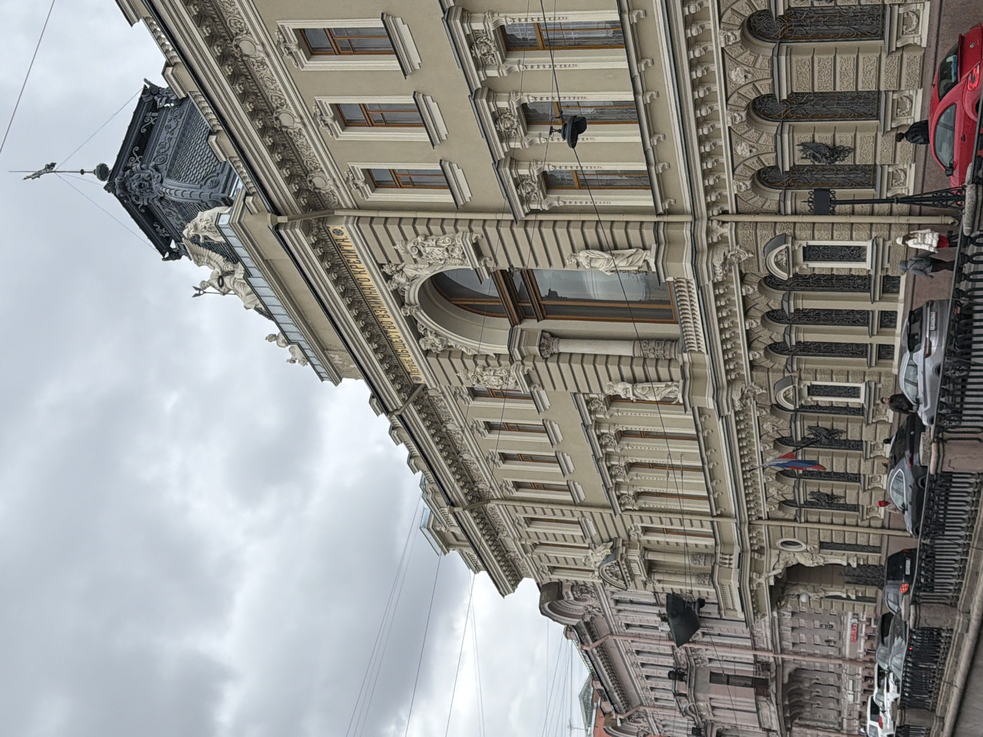 The image depicts the facade of a historic building in Saint Petersburg, Russia, characterized by elaborate architectural details. The building features numerous windows with decorative trim, ornate moldings, statues, and a distinctive tower with a weather vane on top. A golden inscription is visible along the facade, adding to its grandeur. The sky is overcast with heavy clouds.
Along the street, a row of parked cars lines the curb, including red and silver vehicles. Pedestrians are walking on the sidewalk next to the building. A flag is visible hanging from one of the building’s windows. Overhead wires crisscross the sky, adding a modern element to the historic setting.