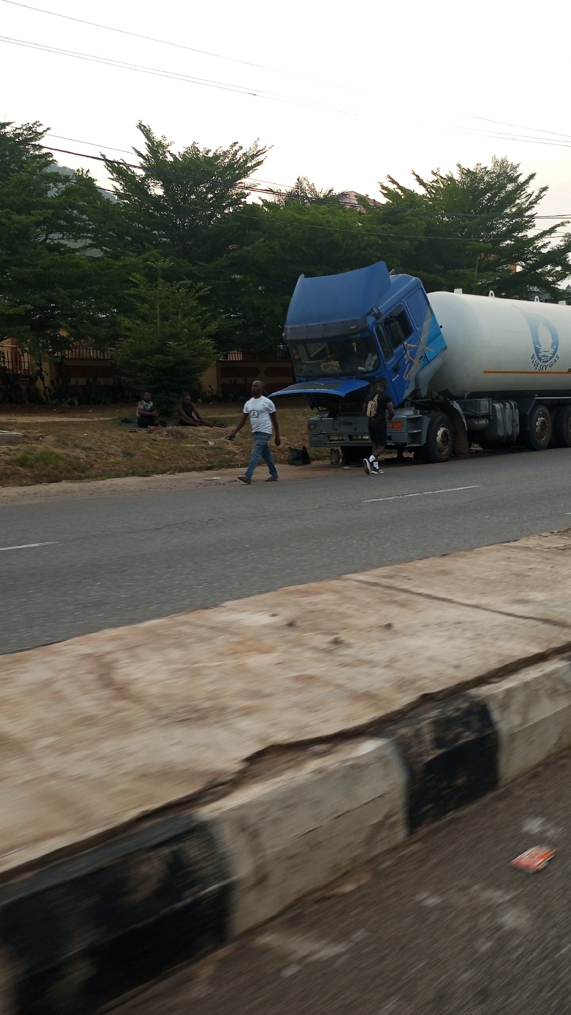 This image captures a roadside scene in Adankolo, Nigeria, featuring a large blue and white tanker truck that appears to be stationary, possibly due to a breakdown or maintenance, as its cab is tilted forward.
Several individuals are visible. In the mid-ground, a man in a white t-shirt, blue jeans, and brown shoes walks from right to left across the dirt verge beside the road. Another man, wearing a black t-shirt, shorts, and a backpack, stands closer to the truck on the right, looking towards the truck's front. Further back on the grassy verge, two or three other men are seated or resting.
The tanker portion of the truck clearly displays the text "BLUE SKY GAS" along with a blue and white logo, possibly representing a flame or a droplet. The truck's cab also shows the numbers "3070" on a small plate.
The scene is set alongside a paved road, which has a concrete curb with black and white stripes in the foreground. Beyond the verge, dense green trees line the background, with faint power lines visible against the bright, possibly overcast, sky. The lighting suggests it could be late afternoon or early evening. The individuals appear calm, with no strong emotions discernible.