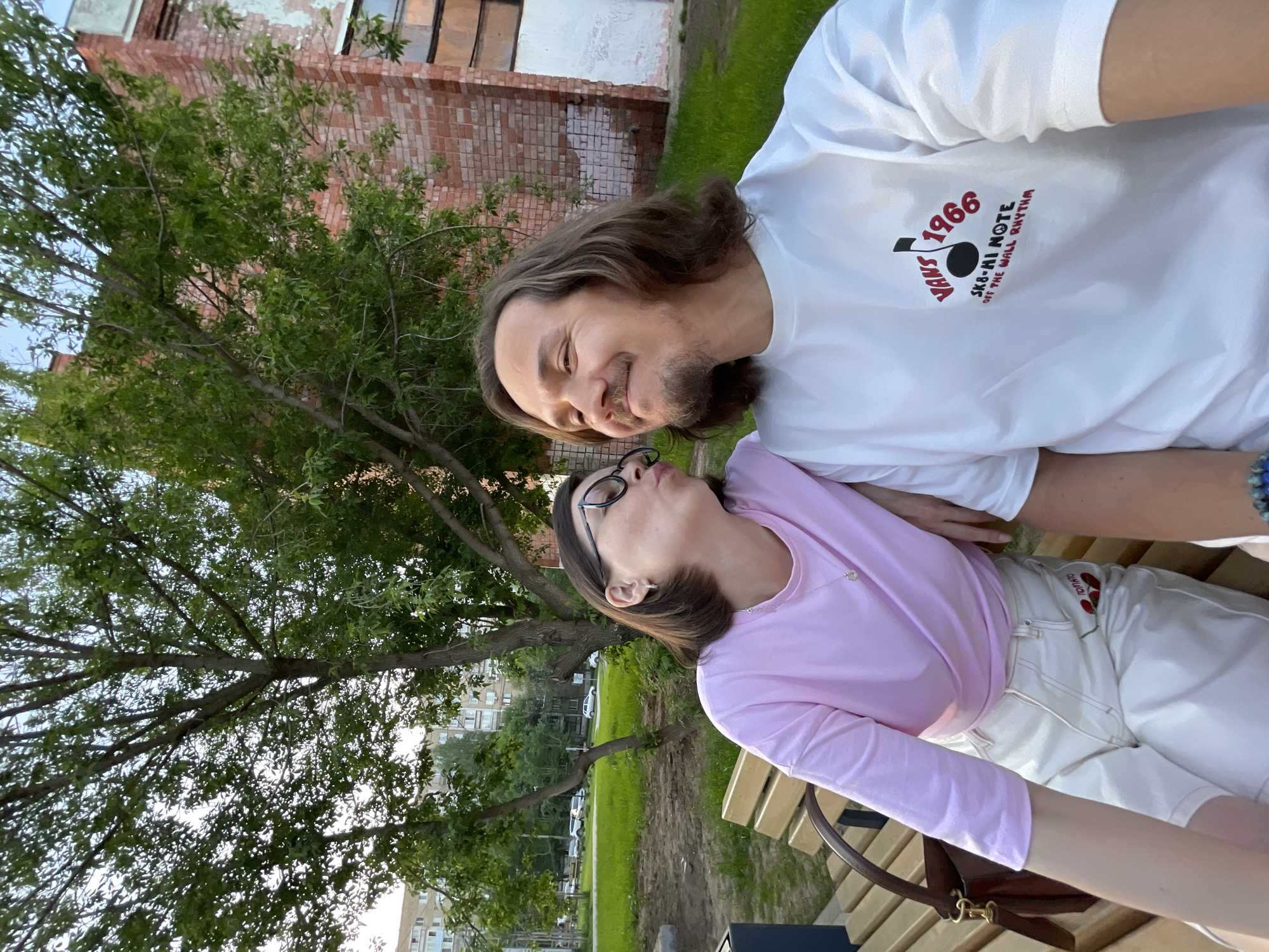 A couple is posing for a selfie outdoors. A man with long brown hair and a beard is smiling at a woman with glasses and short brown hair, who is looking back at him. The man is wearing a white t-shirt with a "Vans 1966" logo. The woman is wearing a pink long-sleeved shirt and white pants. They are sitting on a wooden bench. Behind them is a brick building, grass, and trees. In the background, a residential building can be seen. The location is in Izhevsk, Russia.