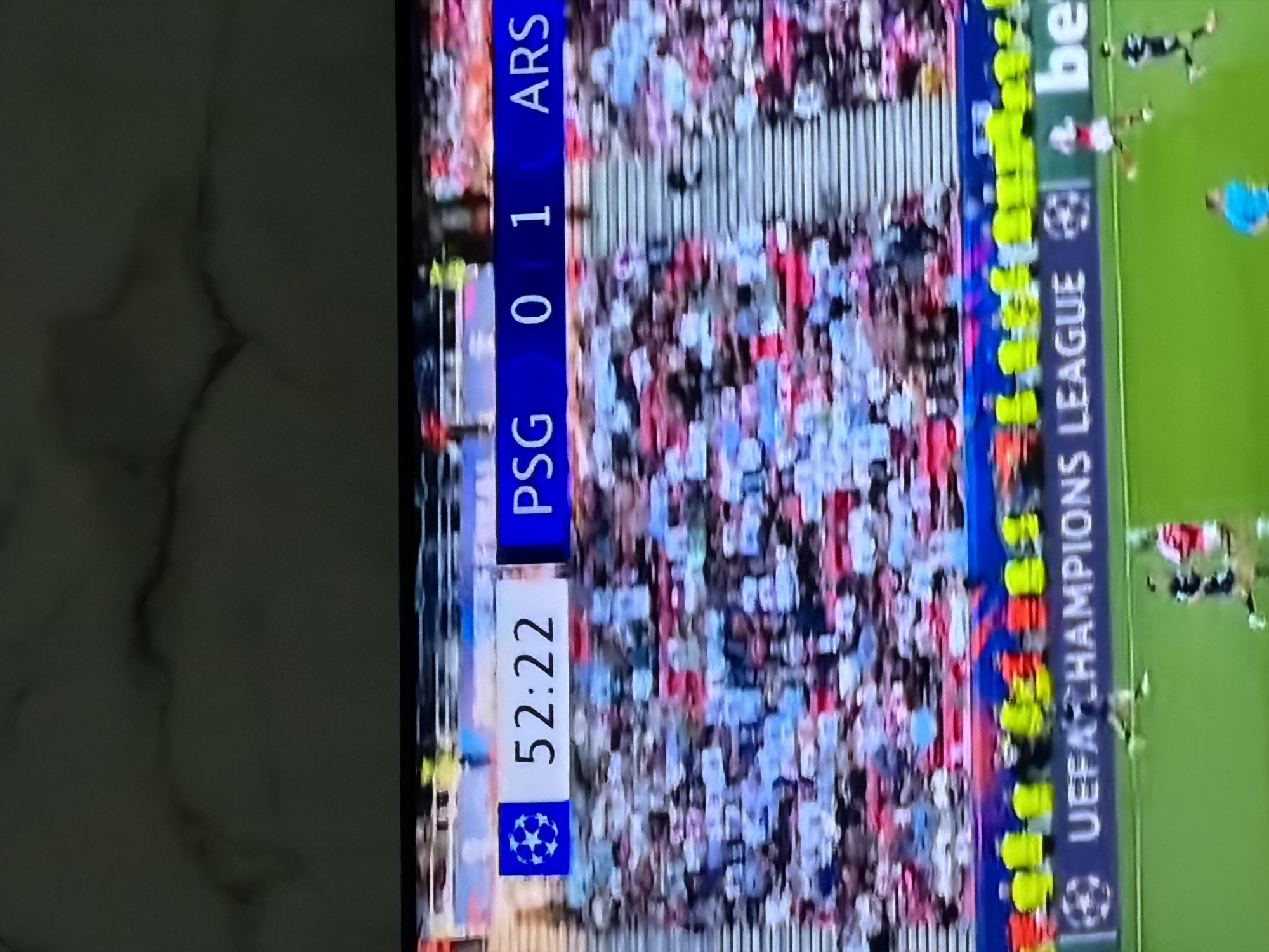 The image shows a television screen displaying a football (soccer) match. The score is PSG 0, ARS 1, with the game clock at 52:22. The match is part of the UEFA Champions League. The background shows a crowd of spectators. No information suggests the image was taken in Asenovgrad, Bulgaria.