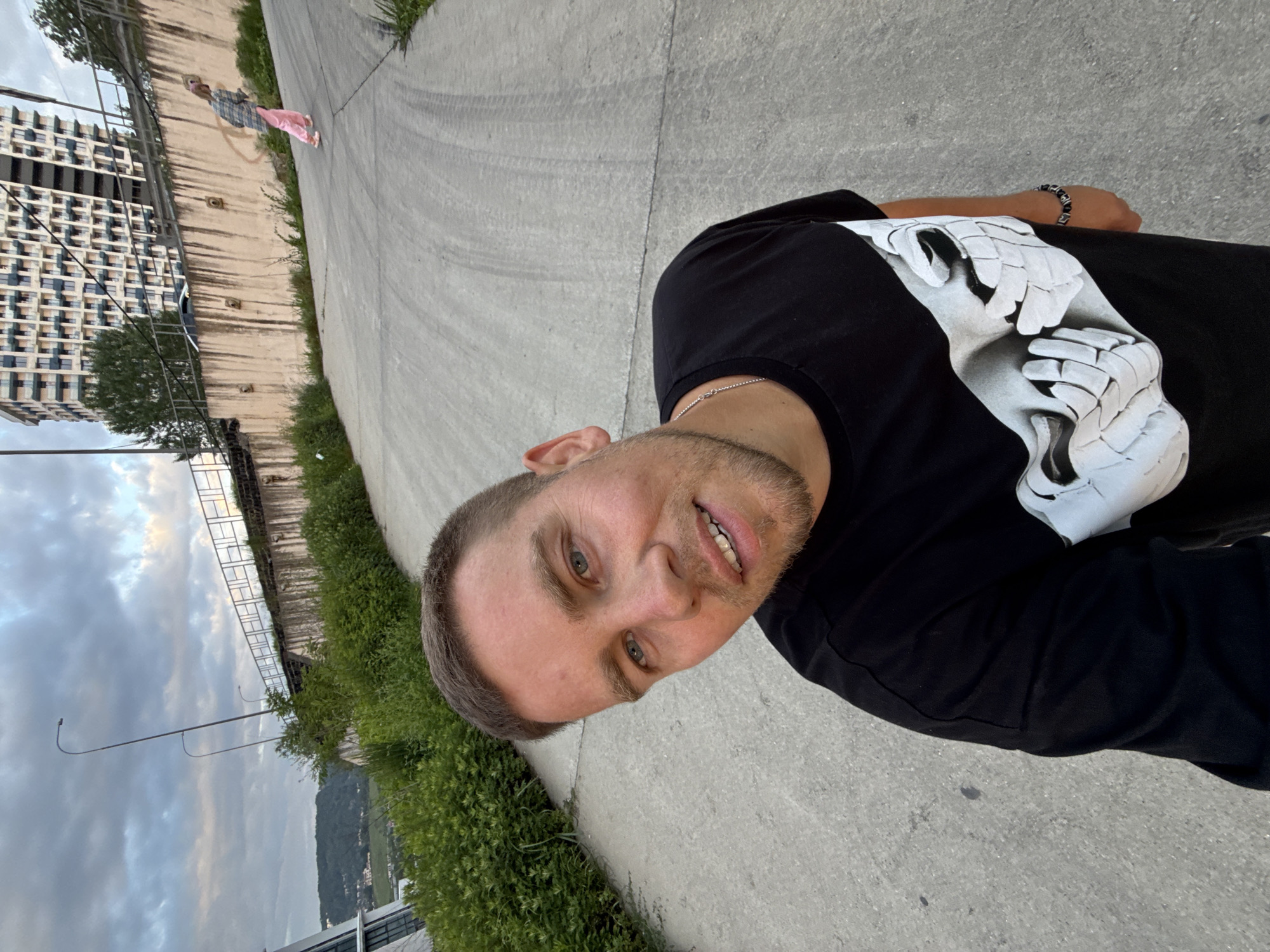A man with short, light-brown hair and blue eyes is posing for a selfie on a concrete walkway. He is wearing a black t-shirt with a white graphic on it. He is wearing a bracelet on his right wrist and a silver chain. In the background, there are buildings and greenery. A person wearing pink clothing is walking on the walkway ahead. The location appears to be in Sochi, Russia, as indicated by the architecture and vegetation.