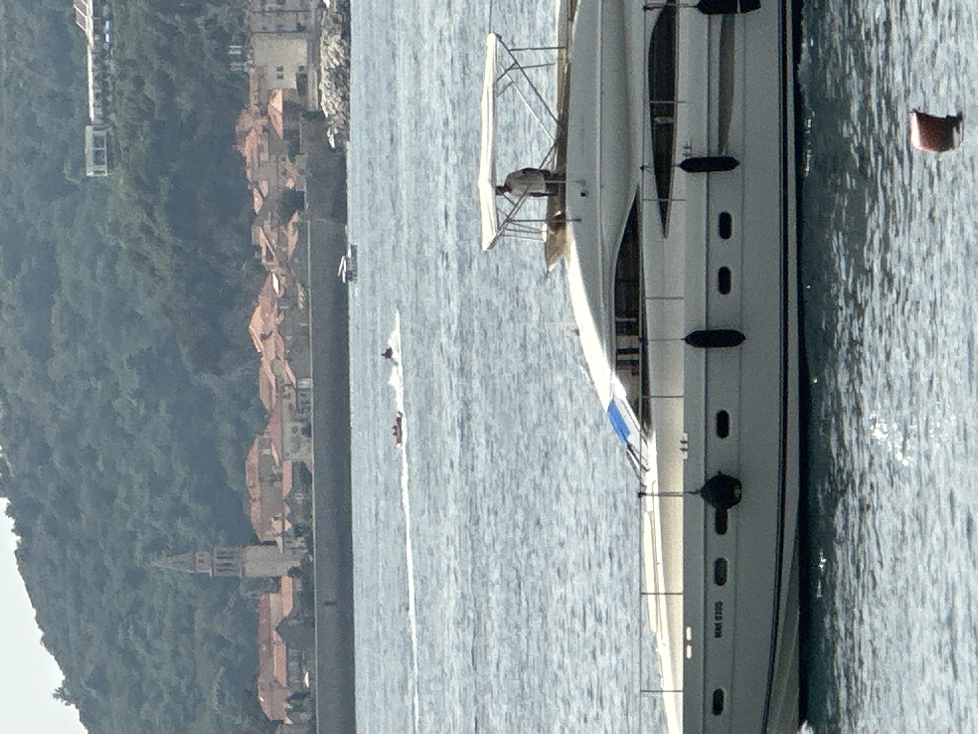 The image captures a sunny day at the Budva Riviera in Montenegro, with the sparkling Adriatic Sea in the foreground and the historic old town of Budva nestled against a lush green hillside in the background. A large, modern white yacht with a Bimini top is prominently featured, its sleek lines cutting through the calm blue water. A single person, wearing a light-colored shirt, stands near the helm of the yacht, appearing to be in control.
Further out in the water, several smaller vessels are visible. A red inflatable towable is being pulled by a speed boat, with two individuals onboard leaving a white wake. In the middle distance, another small boat appears to be cruising. The coastline of Budva is characterized by its charming terracotta-roofed buildings, dense vegetation, and a prominent church tower with a distinctive spire. The overall scene conveys a sense of leisure and the beauty of coastal life in Montenegro.