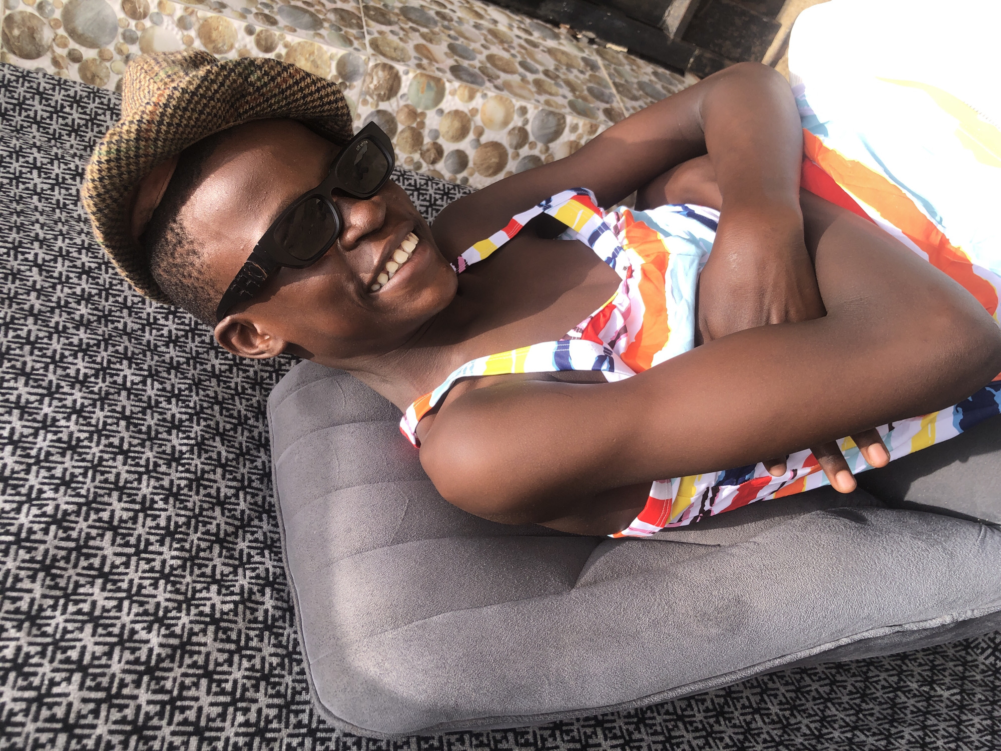 A person with dark skin, wearing sunglasses and a patterned cap, sits with arms crossed in a grey armchair. They are dressed in a colorful, striped outfit. The background features a patterned fabric and a tiled wall. The scene appears to be outdoors, with bright sunlight casting shadows. This image was taken in Ado, Nigeria.