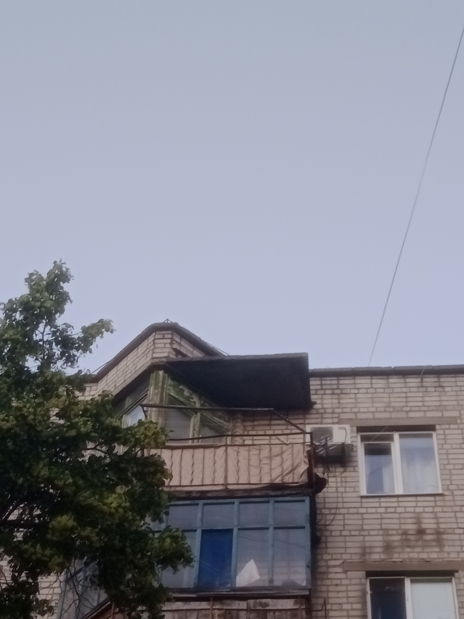 The image shows the facade of a multi-story brick building in the city of Horishni Plavni, Ukraine. The building has several balconies and windows. A tree with green foliage is partially visible on the left side of the image. The sky is overcast. One balcony appears to be enclosed with blue window frames, and another, above it, has a rusted metal railing and a black awning. An air conditioning unit is mounted on the brick wall between two windows on the upper floor. The brickwork is light beige, with some signs of wear and staining. The scene appears to be a residential building in an urban environment.