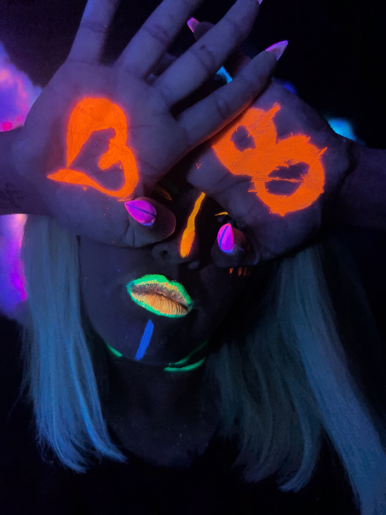This striking portrait features a person adorned with vibrant, fluorescent body and face paint, illuminated by UV light against a dark background, suggesting a blacklight party or an artistic photoshoot. The subject, likely female, has light-colored hair that glows with a blue-cyan luminescence, and her face is partially obscured by her hands.
Her hands are positioned over her eyes, revealing intricate orange fluorescent symbols painted on the palms. On the viewer's left hand, a prominent, stylized orange shape resembles a heart, possibly integrated with an abstract character or a 'B'. On the viewer's right hand, another orange symbol is visible, which could be interpreted as a stylized '8', an infinity symbol, or an abstract double-loop design. Her fingernails are painted with a bright pink and purple fluorescent polish.
The subject's lips are a focal point, painted with fluorescent green around the edges and orange/yellow in the center, creating a vivid, glowing effect. A fluorescent blue line is painted vertically down her chin, with other subtle green lines visible on her neck. The dark setting emphasizes the glowing paint and creates an ethereal and artistic atmosphere, consistent with blacklight-themed events. While the image itself does not provide explicit location cues, it is contextually placed in Noviy, Russia, where such artistic expressions might be found. The time of day appears to be nighttime or an artificially darkened indoor environment, focusing on the luminescent art. The overall impression is one of creativity, expressiveness, and a playful, mysterious mood. There is no discernible textual writing, only abstract symbols.