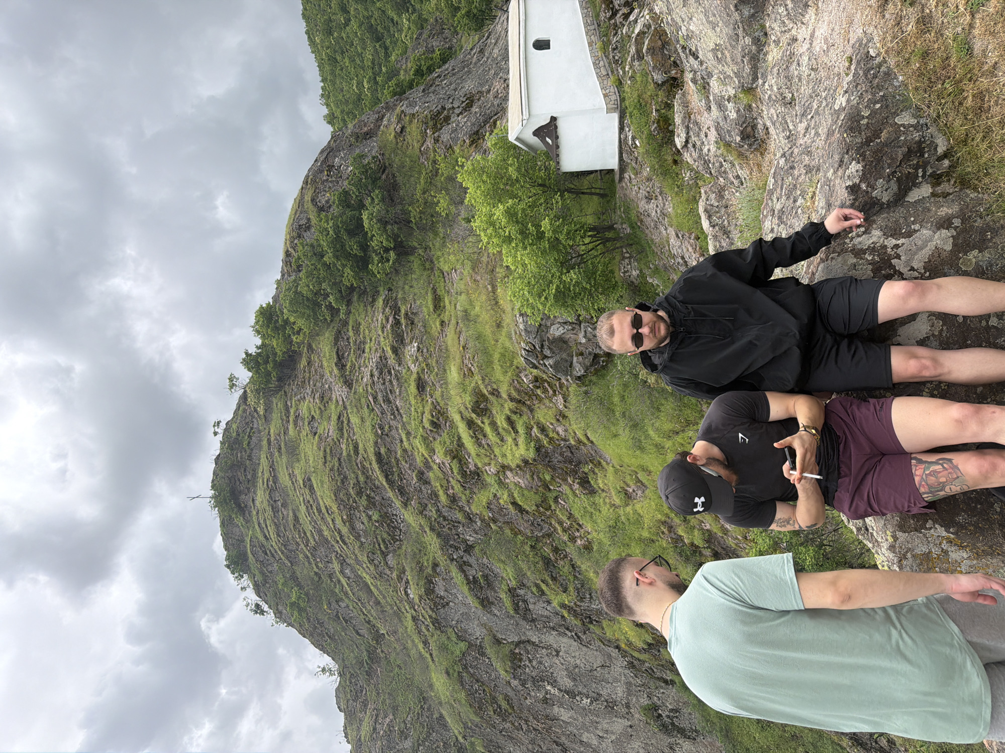 Three men are positioned on a rocky, elevated terrain with a mountainous backdrop. In the background is the Cross Forest church, Ustina, Bulgaria. One man, wearing a black jacket, black shorts, and sunglasses, is lying down. Another man, wearing a black T-shirt, a black baseball cap, and maroon shorts with a tattoo, is sitting and holding a phone and a cigarette. A third man, wearing a light green T-shirt, is standing with his back partially facing the camera. The sky is overcast.