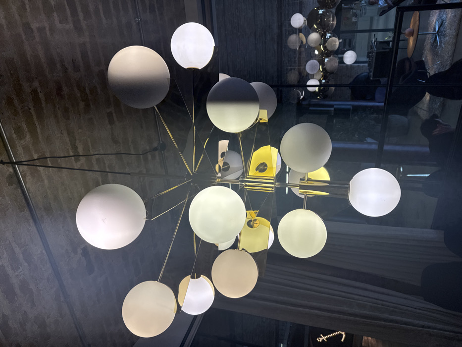 This image captures an interior view of a commercial or hospitality establishment, specifically identified as an area within The Ned hotel, located in the City of London, United Kingdom.
The central subject is a large, modern chandelier suspended from a dark, exposed brick ceiling. The chandelier features a gold-toned metallic frame with numerous radiating arms, each terminating in an opalescent white spherical glass diffuser of varying sizes. Several of these diffusers are illuminated, casting a warm light. Black electrical conduits are visible running across the brick ceiling.
To the left, a light-colored curtain is partially drawn, with a faint, illuminated stylized "f" or similar symbol discernible on the wall behind it. In the bottom left, an indistinct dark shape, possibly a chair or part of a person, is present.
The right side of the image reveals a large glass partition or window, producing a prominent reflection of the primary chandelier. Through this glass, a secondary, similar chandelier is visible in the background, appearing to have reflective, possibly mirrored, spherical elements, some of which are illuminated. Beyond this, a dimly lit lounge or dining area is present, furnished with dark tables and chairs. Indistinct human figures are visible within this background space, suggesting occupancy. A faint reflection of a person's head and shoulder is visible in the bottom right foreground.