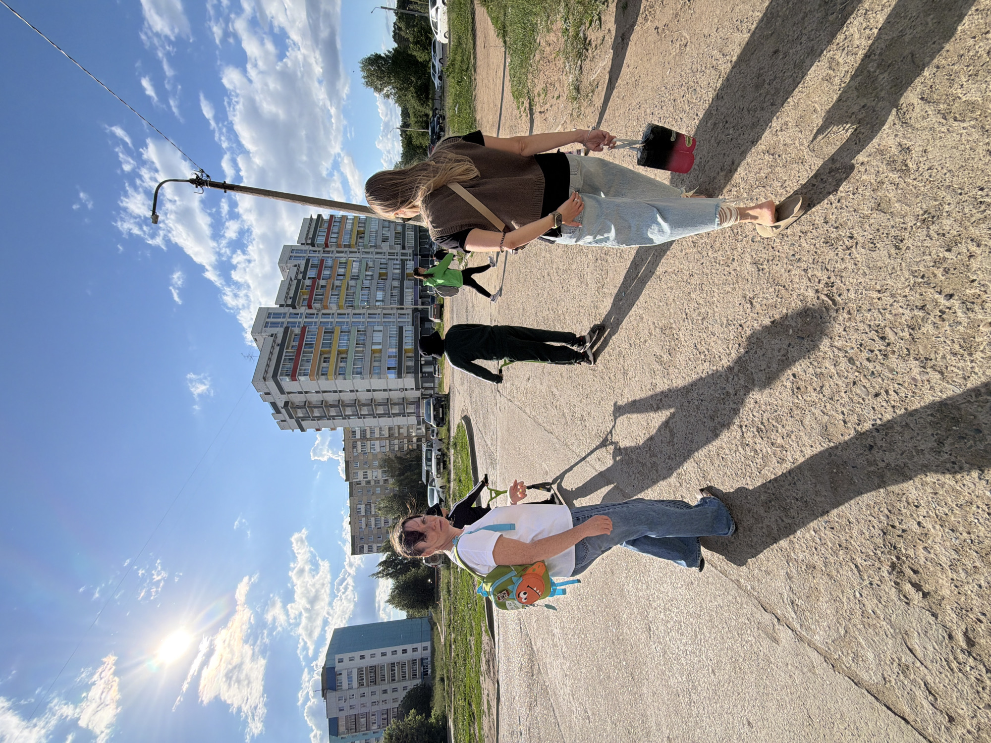 The image depicts an outdoor scene in the city of Chaikovsky, Russia, on a sunny day with scattered clouds. The foreground shows a cracked asphalt and gravel path. Several people are present. In the lower left, a woman with dark hair pulled back, wearing a white t-shirt, blue jeans, and a green backpack with a cartoonish orange monster design, is walking away from the camera, looking over her shoulder. To her right, another person on a scooter is partially visible. Further in the middle ground, a person in a dark green tracksuit and a black hat is riding a scooter. To the right of them, a woman with blonde hair, wearing a dark vest over a black t-shirt, light blue jeans, and sandals, is walking away from the camera, carrying a black and pink cylindrical bag. Another person in a bright green jacket is walking in the background. Several multi-story apartment buildings are visible in the background, some with colorful facades. A street lamp and power lines are also prominent elements of the urban environment. The overall impression is that of a casual daytime activity in a residential area.