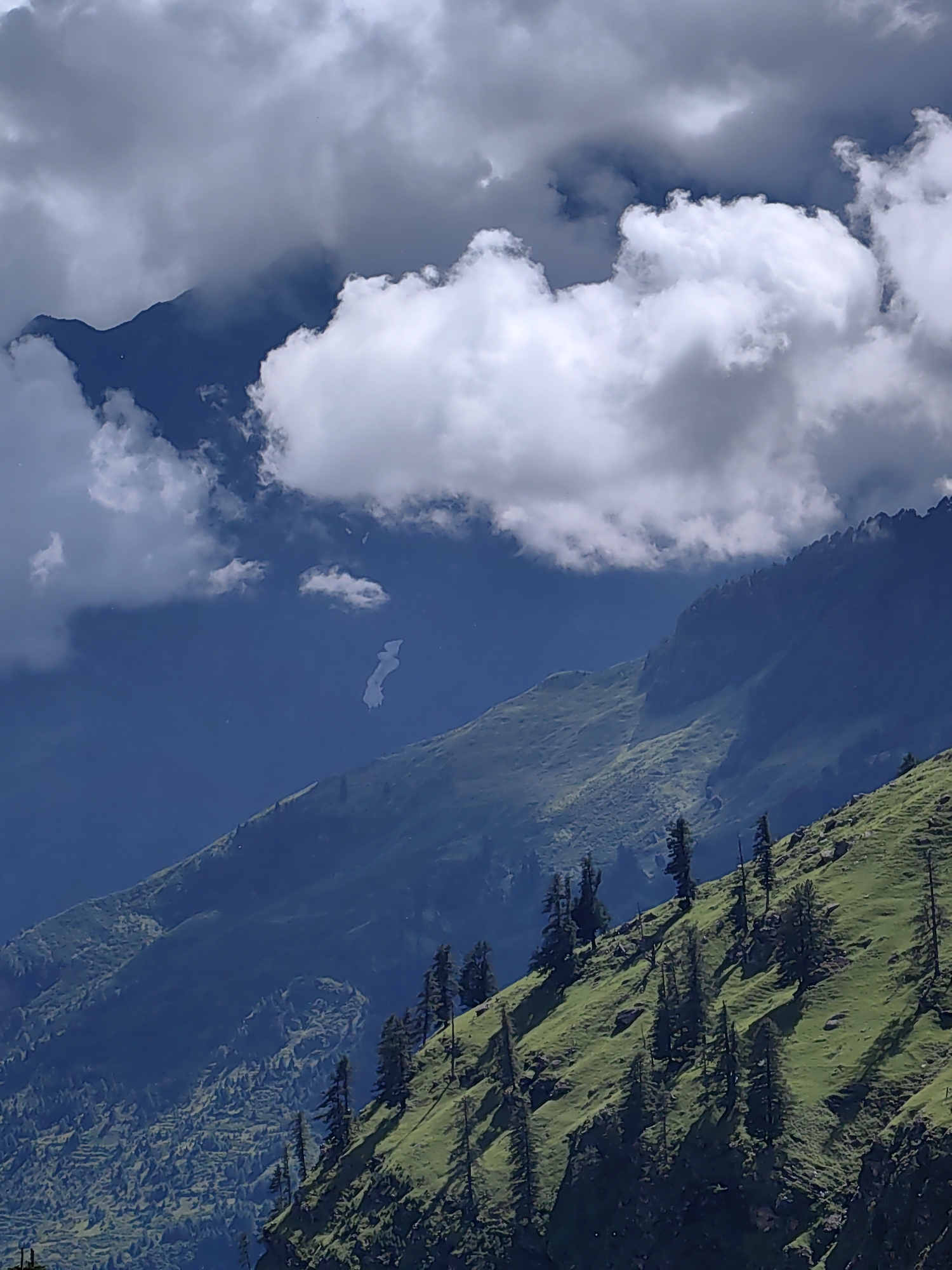 The image shows a view of the mountainous landscape in the vicinity of Kulang, India. The foreground features a steep, verdant slope dotted with scattered coniferous trees, casting long shadows. The middle ground consists of rolling hills covered in lush green grass, with some areas showing terraced cultivation. Further back, darker, more rugged mountain ranges are visible, partially obscured by atmospheric haze and dramatic, fluffy white and grey clouds that dominate the sky. In the distance, a thin, winding line of what appears to be snow or a water body is visible against a deep blue sky. The overall impression is one of a vast, natural, and somewhat remote mountainous region.