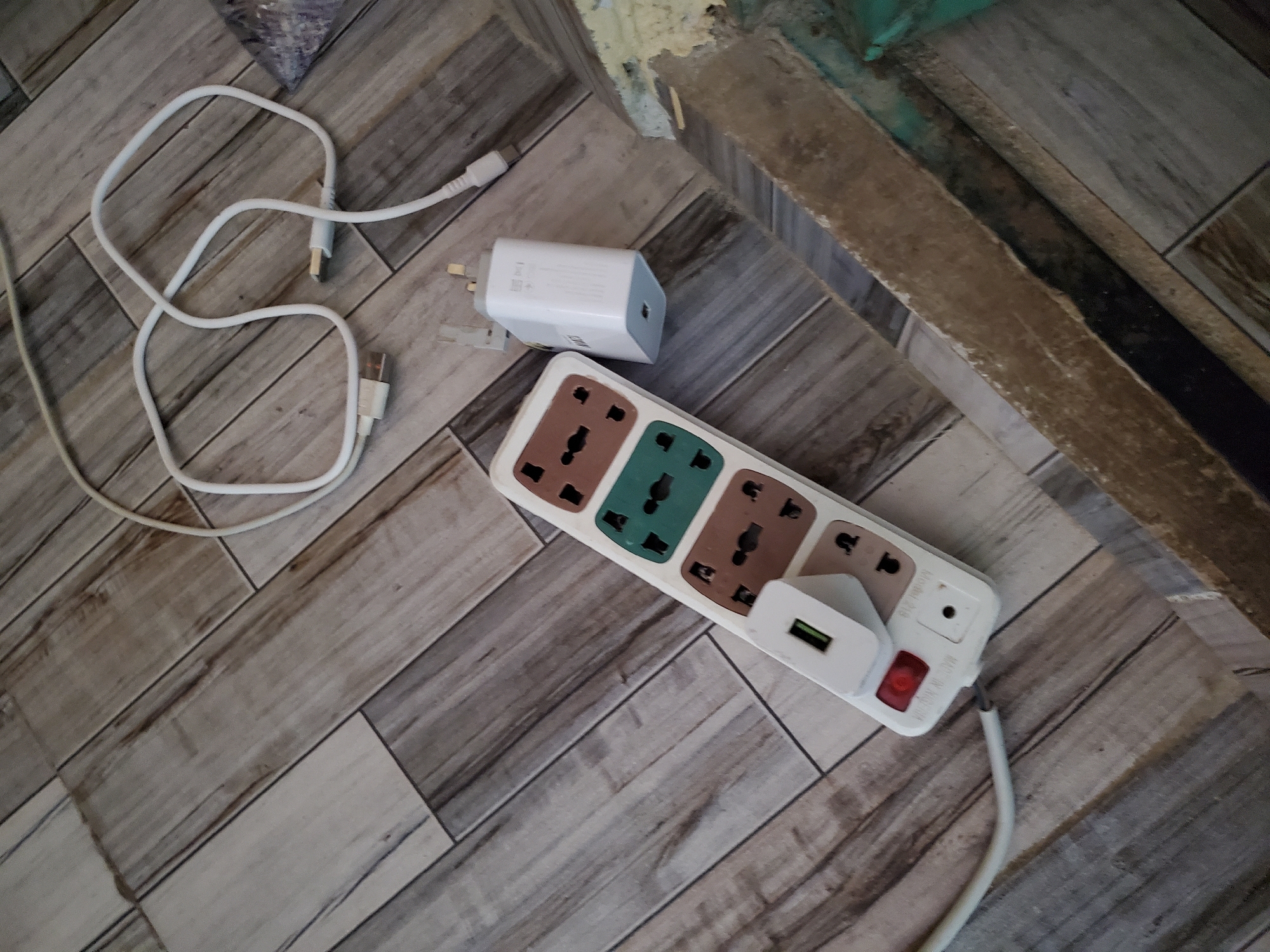The image shows electronic accessories on a wooden floor. A white USB cable is coiled on the floor, with its USB-A connector visible. A white USB charging adapter is next to it. A power strip with multiple universal outlets is also on the floor, with a white USB adapter plugged into one of its sockets. A red power switch is visible on the power strip. The background shows a rustic wooden floor with a texture that resembles aged planks. In the upper right corner, there is a glimpse of a weathered wooden beam and a wall with peeling paint, suggesting an outdoor or unfinished indoor space. The lighting is natural and diffused, indicating daytime. The scene appears to be set in Jalingo, Nigeria, though no specific location cues are visually prominent beyond the general appearance of the flooring and wall. No people are present in the image.