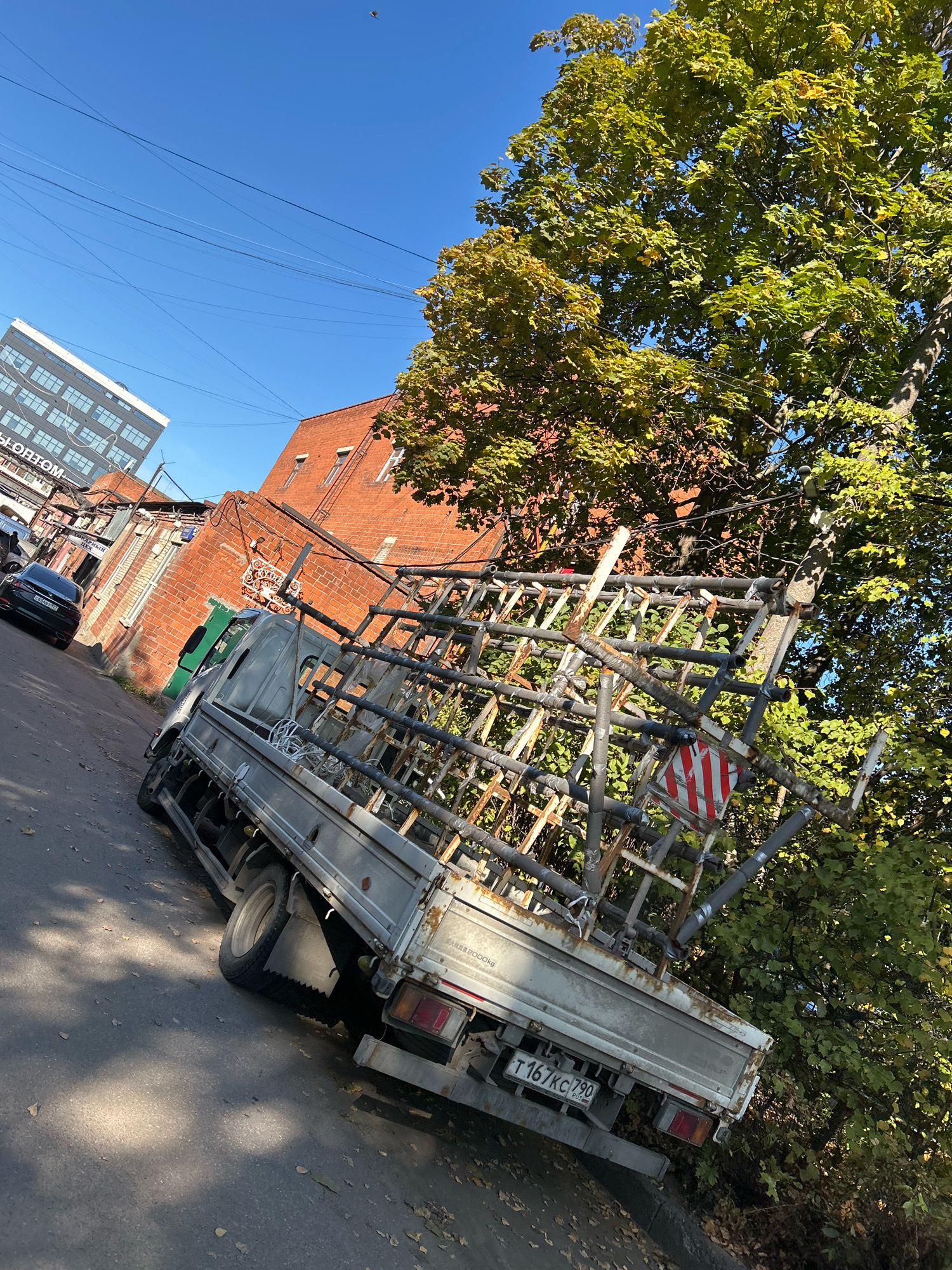 An angled medium shot captures a sunlit street scene in Moscow, Russia. The street slopes downwards, with an asphalt surface showing scattered autumn leaves. On the right, a large tree with green and yellow foliage dominates, its branches casting dappled shadows. Behind the tree, a red brick building rises, its windows suggesting an industrial or older residential structure. To the left, a modern, multi-story white building with numerous windows is visible in the distance, contrasting with the older architecture.
Parked on the street is a light-colored truck, viewed from its rear quarter. The truck has a silver flatbed with a prominent, complex metal frame structure designed for transporting long items, possibly pipes or glass. This frame is constructed from grey pipes of varying diameters, some covered with black insulating material, and appears somewhat weathered. Attached to the frame is a circular sign with red and white diagonal stripes, indicating caution or danger.
Further down the street, a black sedan is parked, facing away from the camera, its license plate partially visible. The sky above is a clear, bright blue, with faint power lines crisscrossing the frame. The overall impression is a typical urban street scene on a clear day, with a focus on the utilitarian truck and its cargo against a backdrop of mixed architectural styles and natural elements.
