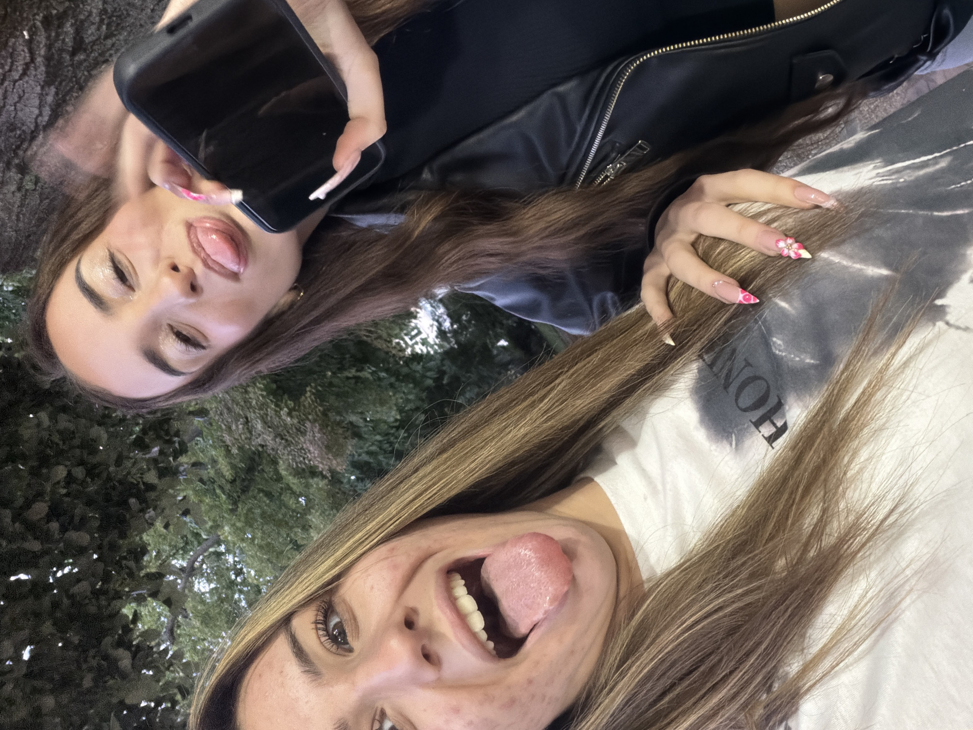 Two young women are posing for a selfie outdoors, with foliage in the background. The woman on the left is closer to the camera and has her mouth open with her tongue sticking out. She has light brown hair and is wearing a white t-shirt with a dark graphic print. She has some acne on her cheek. The woman on the right is holding a black smartphone in her right hand, also sticking her tongue out. She has long, dark brown hair and is wearing a black leather jacket over a black top. Her fingernails are decorated with pink and white floral designs. The lighting suggests it is evening or overcast.