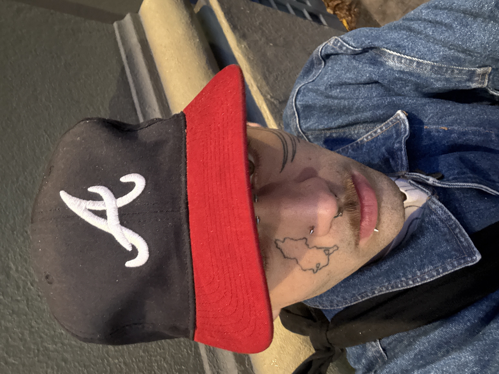 A person wearing a black baseball cap with a white "A" logo and a red bill, a denim jacket, and a black shirt, is looking at the camera. The person has multiple facial piercings and tattoos. The background appears to be an outdoor urban environment with stone steps. There is no information to infer that the location of the image is in the city of Buenos Aires, Argentina.