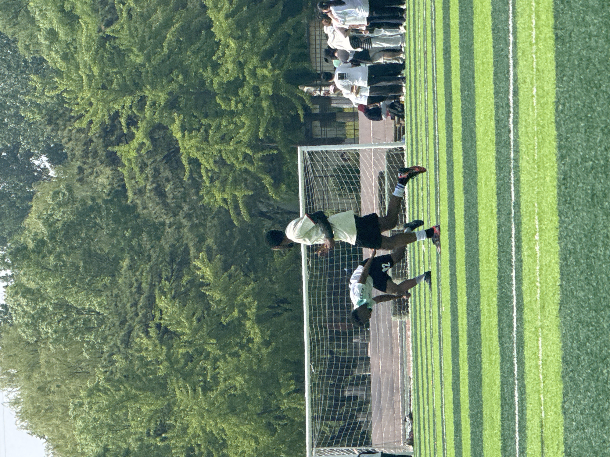 A photograph depicts two male individuals playing soccer near Linghe, China. One individual is in mid-air, near the goal, while the other player is nearby. The scene takes place on a striped green soccer field, surrounded by trees. A group of people are seen on the sidelines in the background.