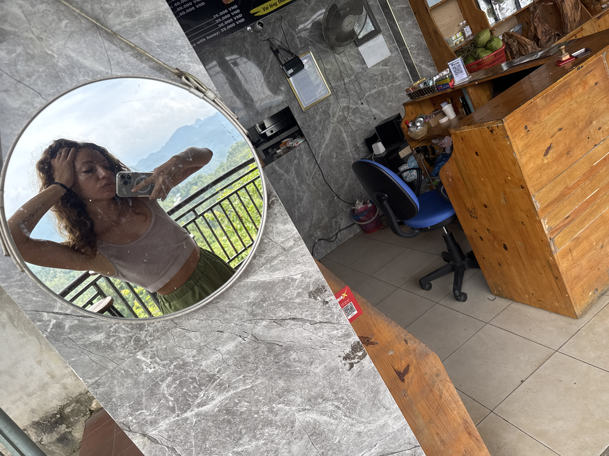 This image captures a woman's selfie in a mirror, against the backdrop of what appears to be a shop interior, likely located in Quản Bạ Commune, Vietnam. The woman is holding her phone, posing in front of a round mirror. The reflection shows her wearing a tank top and green shorts, with a view of mountainous terrain and green vegetation outside.
The shop interior features a grey marble-like countertop where the mirror is placed. In the background, there is a wooden counter with items like coconuts and other goods on display. A blue chair is situated near the counter. The walls are a grey textured material, adorned with a wall-mounted fan, a framed document, and a menu board with Vietnamese text and prices. The floor is tiled, and there is a wooden structure beside the countertop. A sticker with a QR code is visible on the wooden structure.
