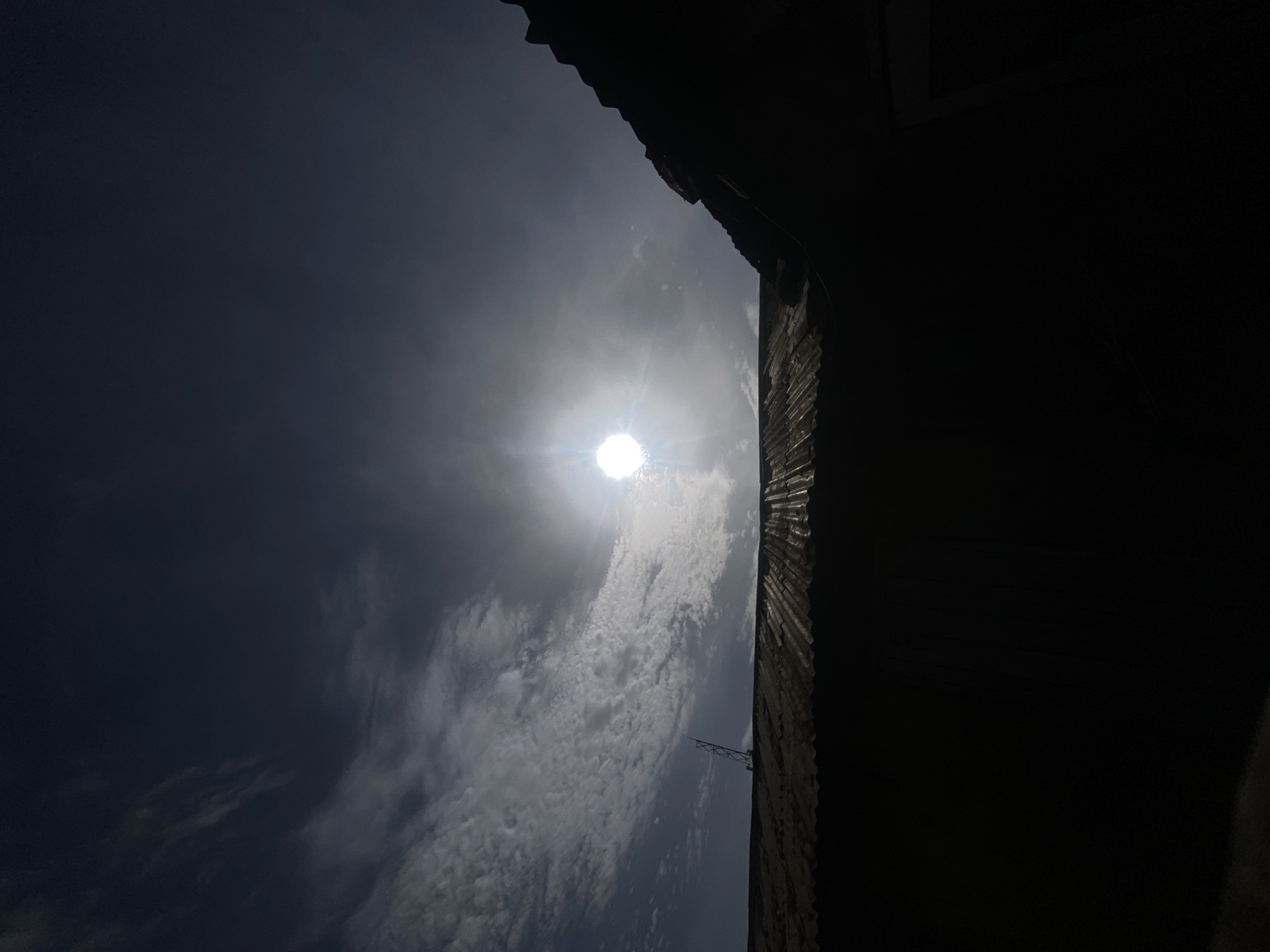 The image shows a view of the sky in Barden Yana, Nigeria. The sun is visible through thin clouds, creating a bright glare. To the right of the sky, part of a dark structure, potentially a building with corrugated iron sheets, is visible. An antenna is attached to this structure. The sky transitions from dark to light with cloud cover.