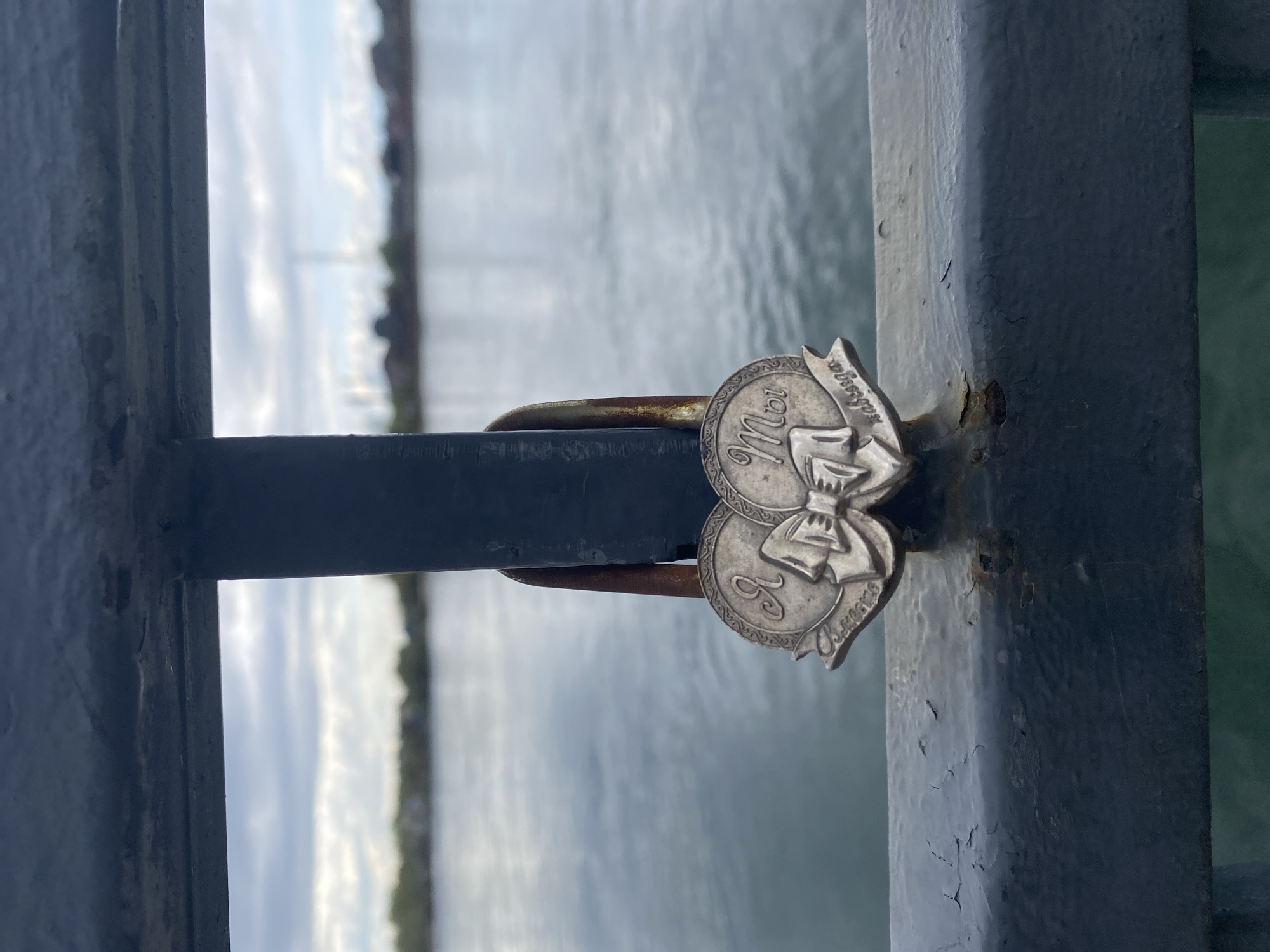 A silver-colored, decorative padlock is secured to a dark-colored, weathered metal fence. The padlock consists of two circular medallions, each with inscribed text in what appears to be Russian Cyrillic. There is a decorative bow connecting the two medallions. The fence overlooks a body of water, partially visible through the fence's openings. The background suggests a partially cloudy sky and some distant shoreline vegetation. The scene is possibly Irkutsk, Russia, based on the Cyrillic inscription on the lock.