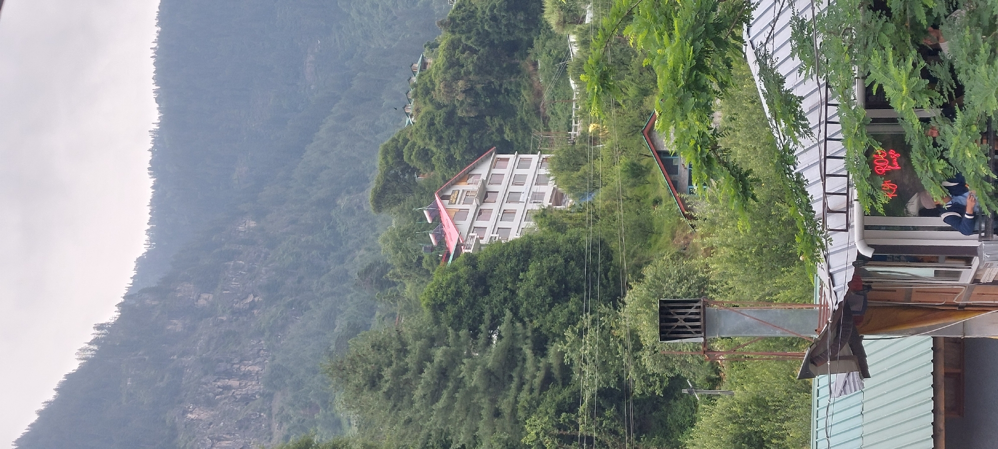 In Vashisht, India, a large, multi-story hotel with a red roof is nestled on a hillside amidst lush green trees and a rocky mountain in the background. The hotel, likely a resort given its scenic location, features a white facade with numerous windows. Below the hotel, other buildings, including a smaller stone structure with a green roof and a shop with a corrugated metal roof, are visible. Several power lines crisscross the scene. In the foreground, a metal exhaust pipe or chimney stands tall, partially obscuring a shop with a neon sign that reads "Ky ROO house." Two individuals are partially visible inside the shop. The overall impression is a remote, mountainous landscape with visible signs of human habitation and tourism.