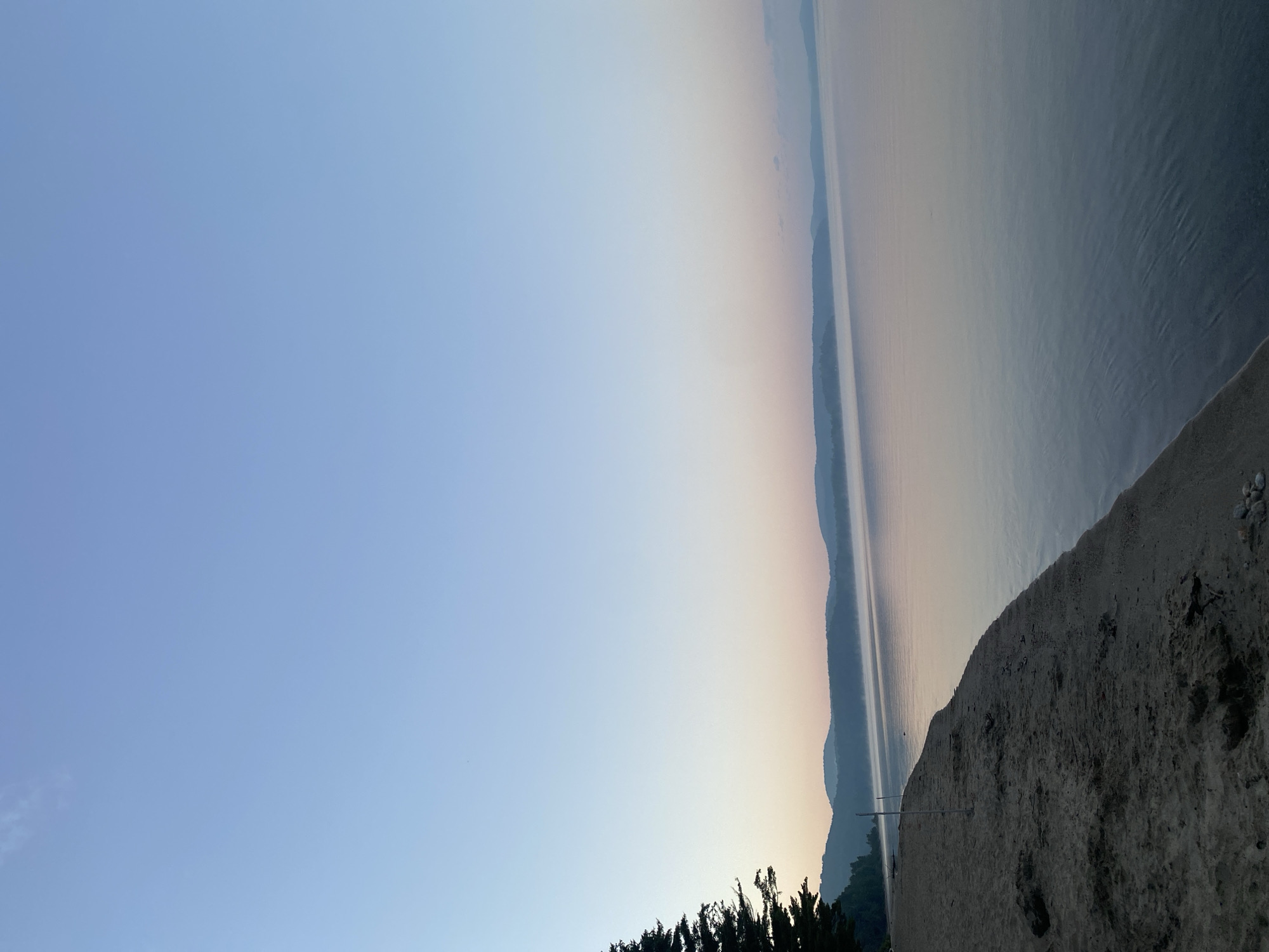 The image depicts a serene lake scene at dawn or dusk, with a sandy shoreline in the foreground. A large evergreen tree stands on the left side of the beach. The water is calm, reflecting the soft hues of the sky, which transitions from a pale blue at the top to shades of pink and purple near the horizon. Distant hills, covered in a light mist, form a hazy silhouette across the landscape. Two wooden posts are visible on the sandy beach, one closer to the water and another further back. The environment appears tranquil and natural, with no signs of human activity or urban presence. The image is taken from a slightly elevated perspective, looking out over the lake.