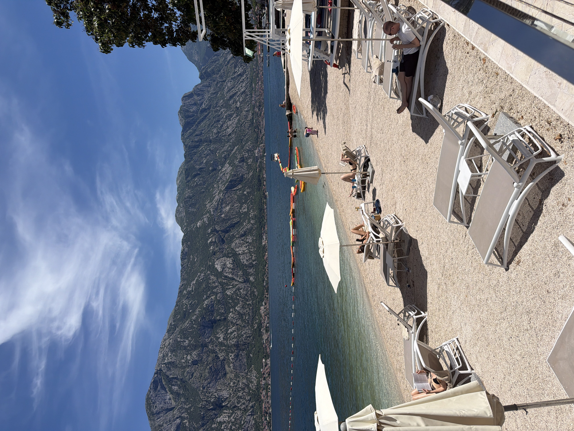A beach scene at Prčanj, Montenegro features a beach with white lounge chairs and parasols on the right, overlooking a body of water and a large mountain on the left. People are relaxing on lounge chairs on the beach. A string of colorful inflatables and buoys marks an area in the water. The sky is blue with scattered clouds. A rocky mountain occupies the left side of the image.