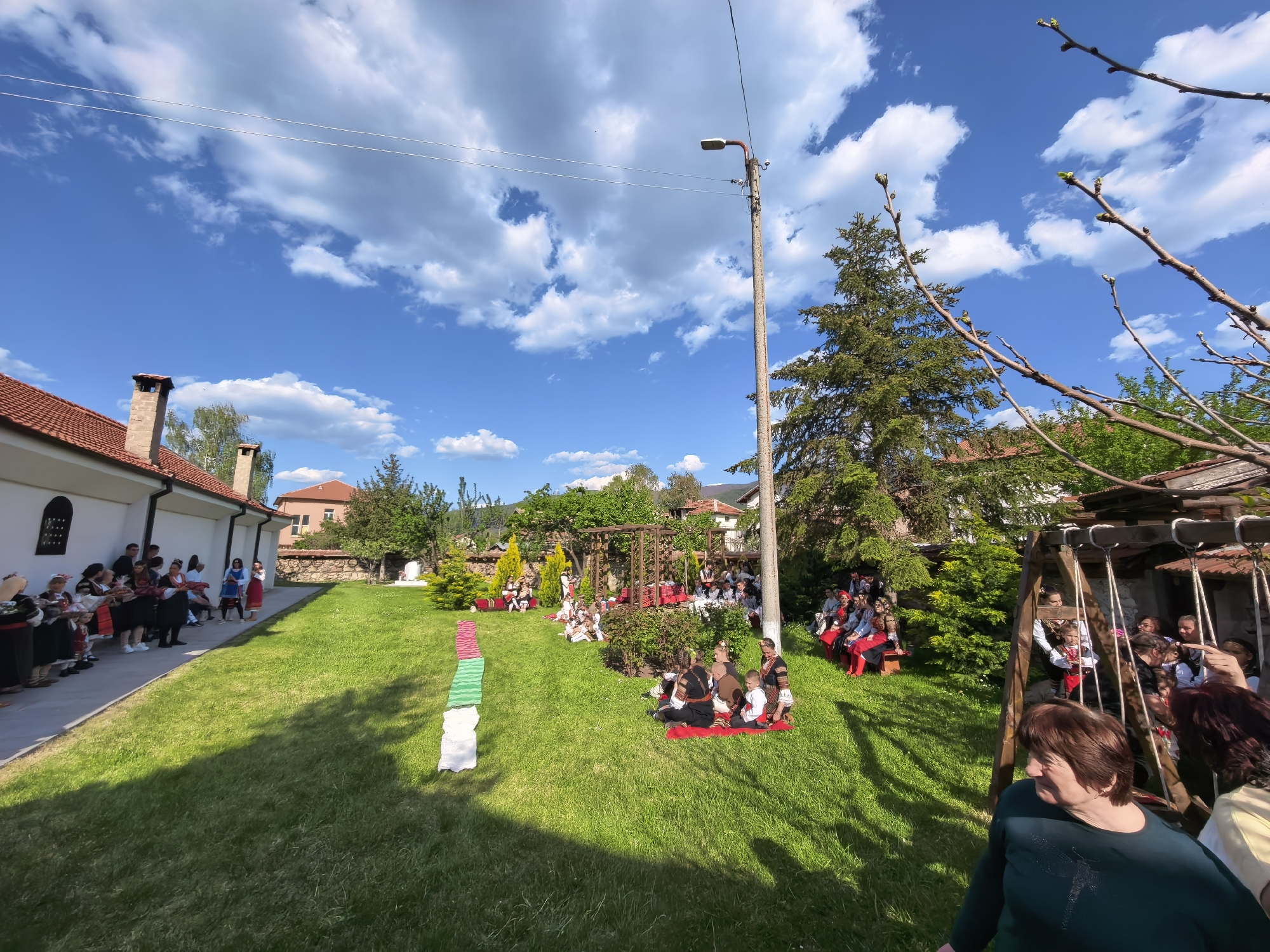 The image depicts an outdoor scene in Sapareva Banya, Bulgaria. People are gathered on a grassy lawn, some seated on rugs and benches. Some are dressed in traditional Bulgarian clothing. A white building is located on the left with people in formal wear. A tall light pole stands near a large evergreen tree. A wooden structure resembling a swing set is on the right. In the foreground, a woman in a dark green shirt is partially visible. The sky is blue with scattered white clouds. Green, white and red fabrics are laid out on the lawn, resembling the Bulgarian flag.