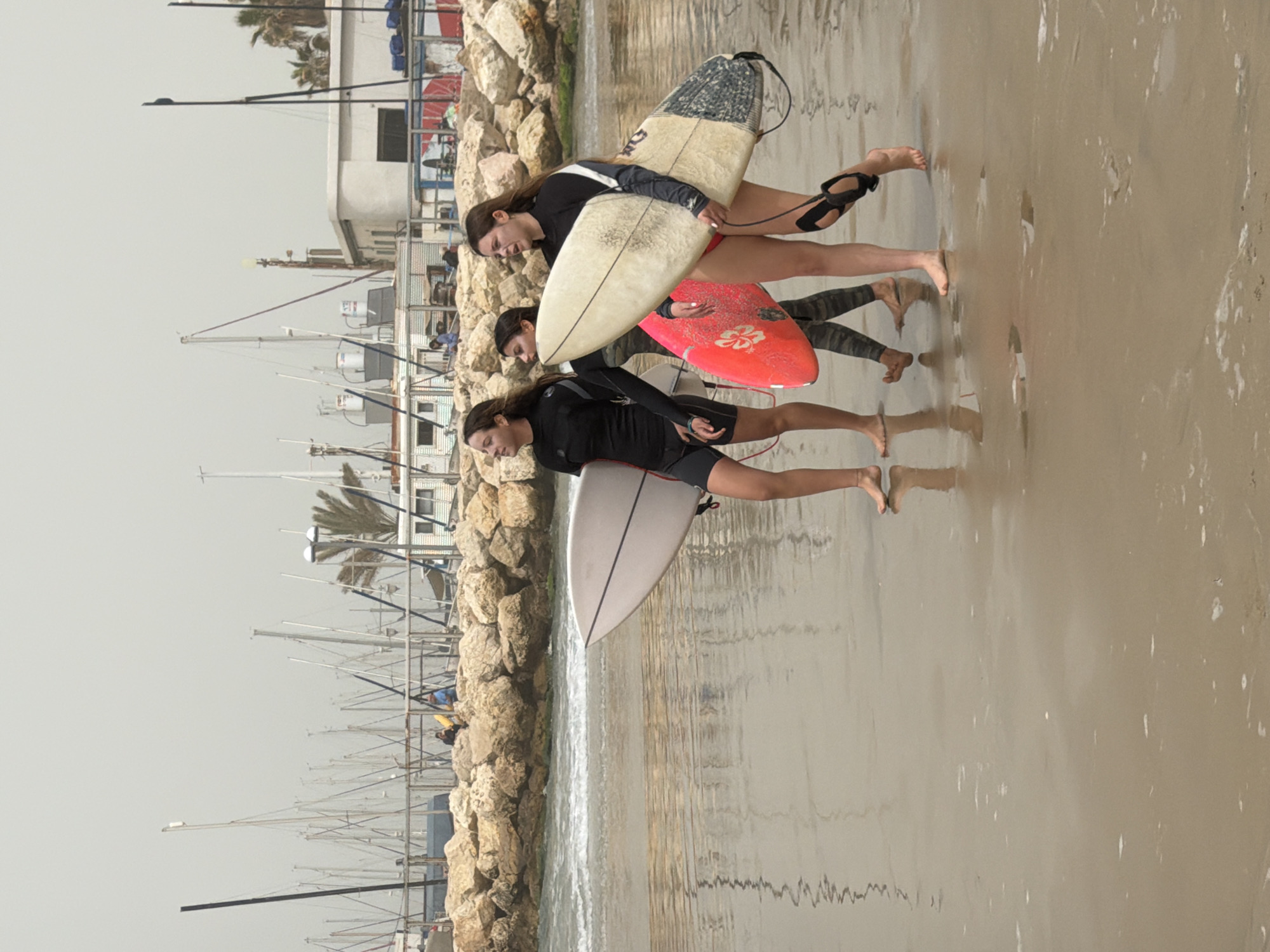 Three young women are walking on the beach in Tel-Aviv, Israel, each carrying a surfboard or bodyboard. The first woman is carrying a surfboard that is mostly white but has some dark patches. The second woman has a red bodyboard with a white flower design. The third woman is carrying a white surfboard with black lines. The women are walking on the wet sand near the water's edge. To the left of the women are rocks and then a harbor filled with many small boats, the masts rising to the sky. The sky is overcast.