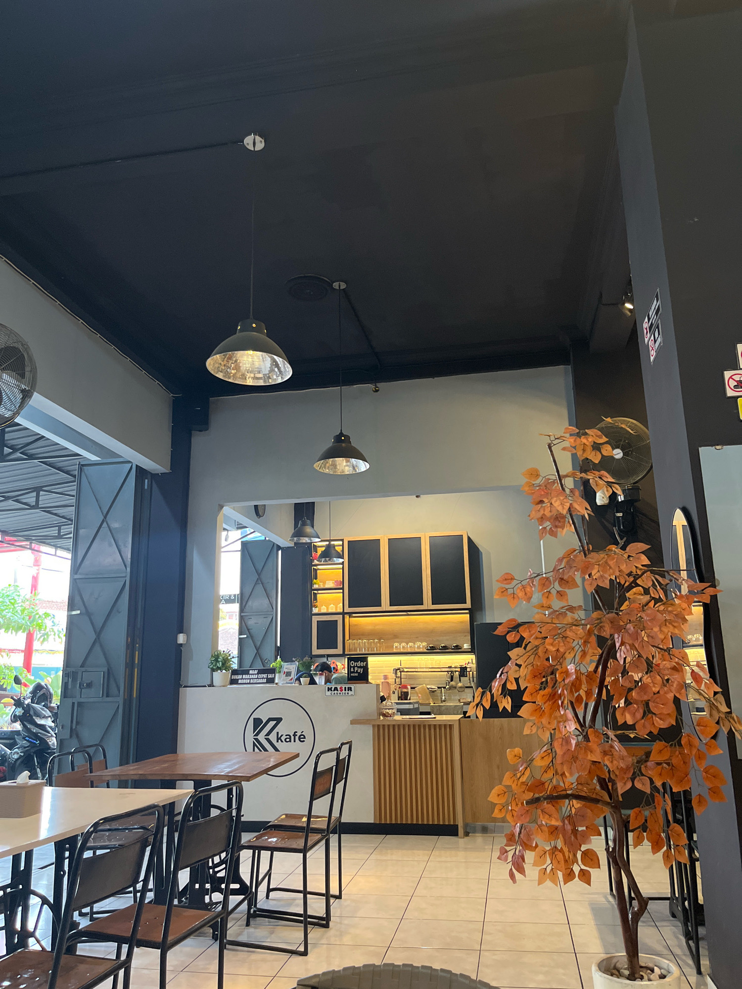 The image shows the interior of a cafe. In the foreground, there are several tables and chairs. To the left, there are large open doors leading outside, revealing a street scene with a motorcycle. In the midground, there is a counter with a coffee machine and shelves stocked with glasses. A decorative tree with orange leaves is positioned to the right of the counter. The cafe has a modern industrial aesthetic with black ceilings and grey walls. The logo of "K kafe" is visible on the counter. The flooring appears to be tiled. The scene is brightly lit, likely by artificial lighting from overhead pendant lamps and spotlights on the shelves. The overall impression is that of a contemporary, casual dining establishment.