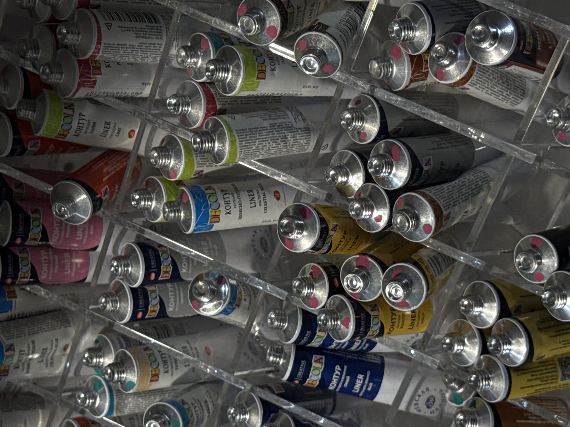 The image displays a close-up view of numerous tubes of "DECOLA KONTYR" (liner) art supplies, arranged in clear plastic holders. The tubes are of various colors, including white, yellow, blue, and pink, and are labeled with Russian text. Some tubes are partially obscured by others. The lighting suggests an indoor setting, possibly a retail display or storage area. No people, actions, or interactions are visible. The location is identified as Moscow, Russia.