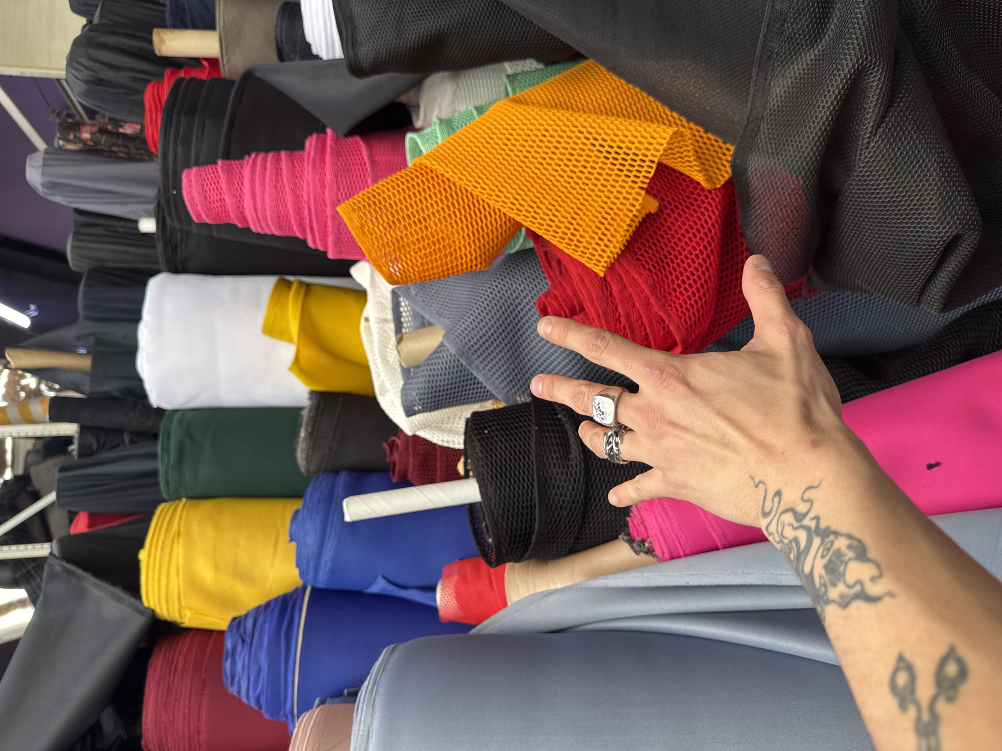 The image depicts a close-up view of a fabric store, likely in Sofia, Bulgaria, given the context. The foreground is dominated by a person's right hand, adorned with two rings and a tattoo on the wrist. The hand is positioned as if selecting or interacting with the surrounding fabrics.
The environment is a vibrant and chaotic display of various rolls of fabric. Dominant colors include bright red, yellow, dark blue, a muted blue-grey, hot pink, green, orange, and black. Textures range from smooth and tightly woven to netted and mesh-like materials. Some fabrics are neatly rolled, while others are draped or bunched, creating a dynamic visual. The background shows more rolls of fabric stacked high, suggesting a large inventory. The lighting appears to be artificial, typical of an indoor retail space. There are no other people clearly visible in the frame.