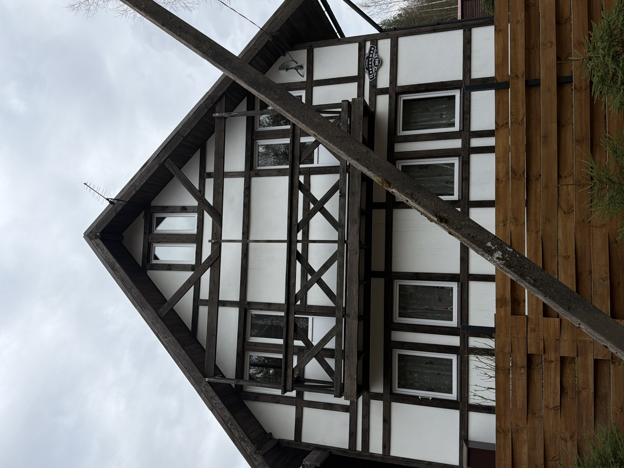 The image shows a two-story building with a distinctive half-timbered facade, characterized by dark brown wooden beams forming a grid pattern on a white background. The building has a steeply pitched roof with a sharp gable end visible on the left. A sign with Russian text and the number 982 is affixed to the upper part of the facade. Several windows with white frames are visible. A wooden fence runs along the right side of the building. A diagonal wooden beam obscures part of the view. The location appears to be Glubokoe, Russia, based on the signage. The sky is overcast.
