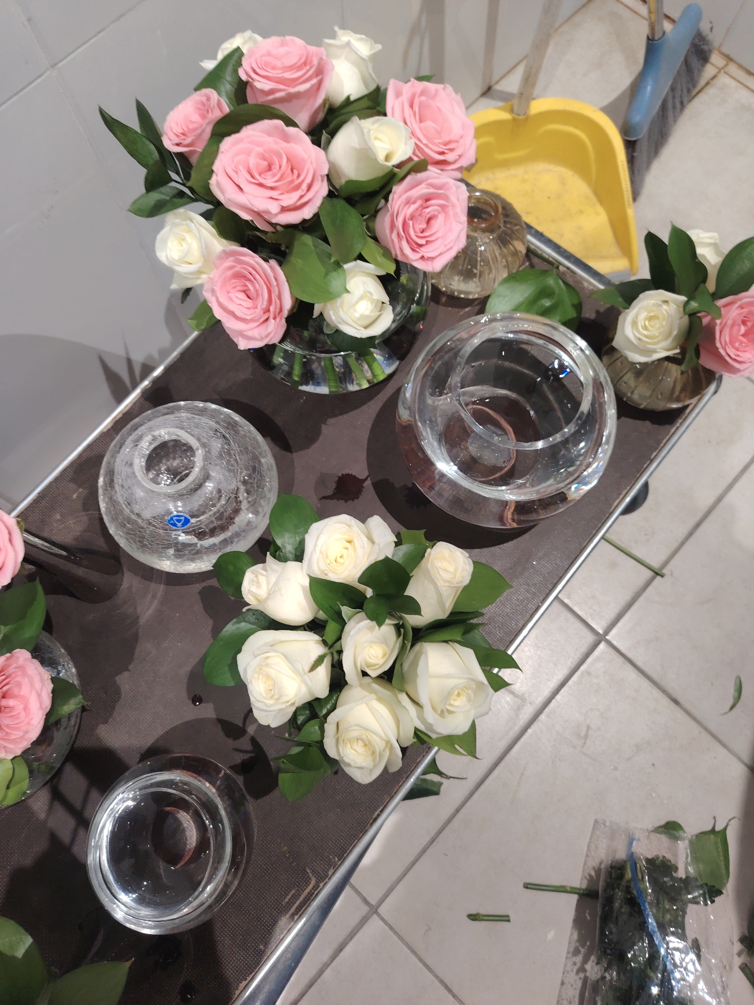 A bouquet of pink and white roses is arranged in a glass vase on a table. Beside the vase, a yellow dustpan and a blue broom are leaning against a wall. The scene appears to be indoors, with a tiled floor and a gray tiled wall. There are no people or animals visible in the image. The overall impression is of a floral arrangement being prepared or displayed. The image does not provide any information about the city of Konigil, Uzbekistan.