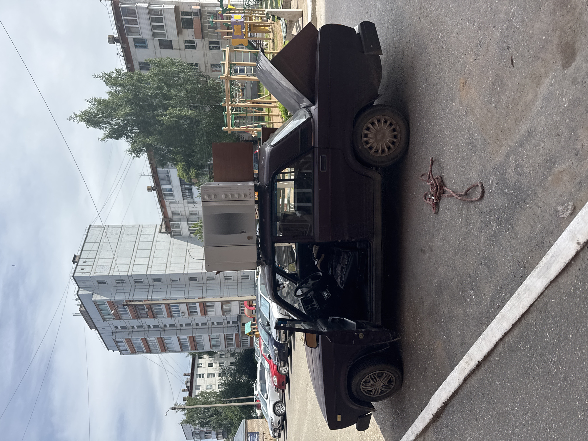 The image captures an outdoor scene in a residential area of Chaikovsky, Russia, during daytime under a partly cloudy sky. The main subject is an older, dark-colored sedan, likely a Lada, parked on asphalt pavement. The car has its hood open and the driver's side door ajar, revealing the interior, including the steering wheel. On the car's roof, a white appliance, possibly a washing machine, is placed or strapped, alongside a flat, brown panel or piece of furniture. The car's trunk is also open, showing items inside. On the ground near the car's rear, a coiled, reddish-brown rope or chain is visible.
In the background, several multi-story apartment buildings, characteristic of Soviet-era architecture, are prominent with visible balconies and windows. A large green tree stands between the buildings and the parked vehicles. A wooden playground structure with colorful elements is visible behind the car. A line of other parked cars, including red and white models, extends to the left. The asphalt ground features a white painted line, possibly marking a parking space or road edge. A partially obscured sign in the lower left background appears to display the Russian word "РЕМОНТ" (REPAIR). The scene suggests an activity of loading or transporting household items in the car.