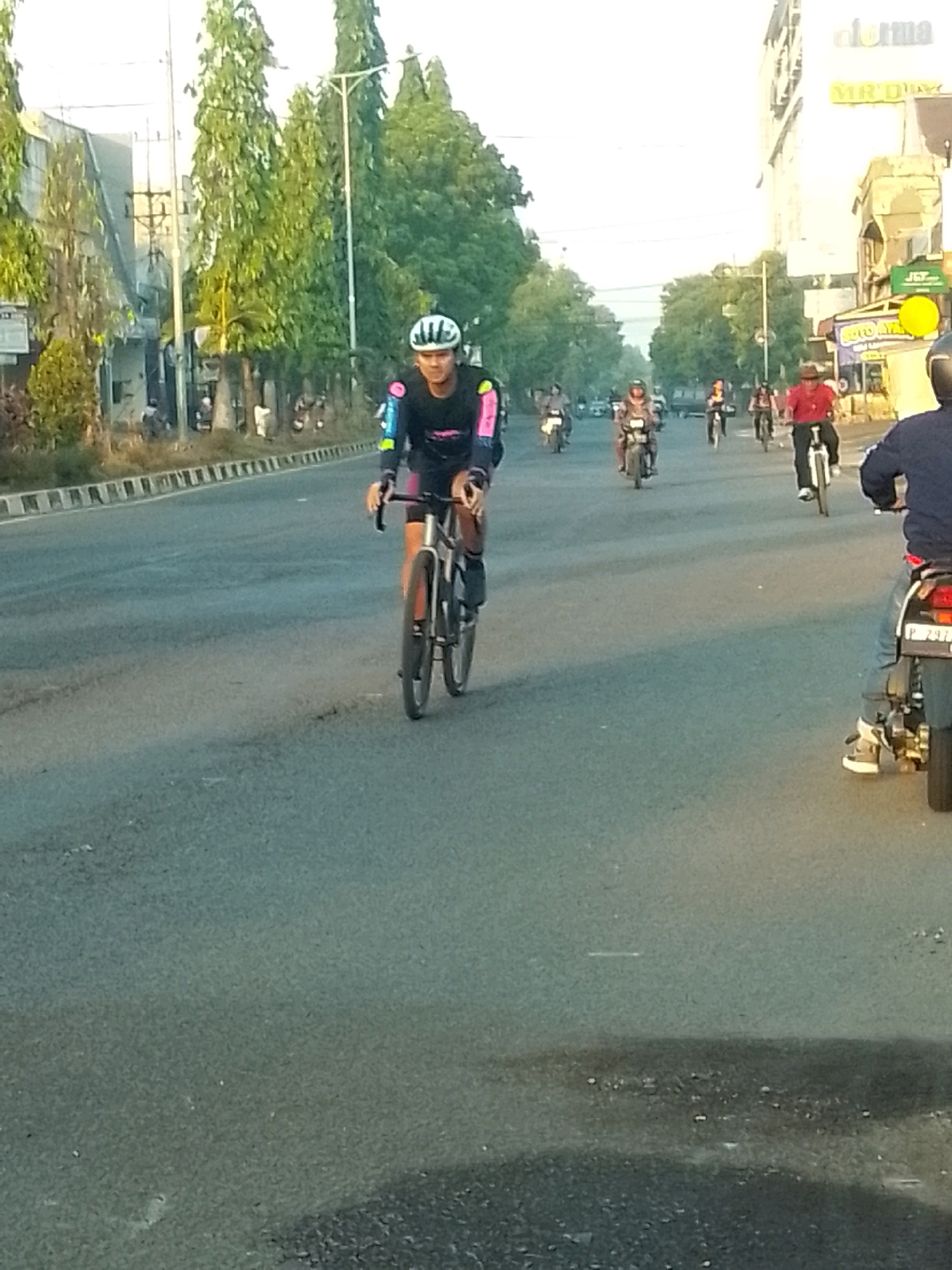 A cyclist wearing a white helmet and a black and pink long-sleeved shirt rides a bicycle on a paved road in Jember, Indonesia. The cyclist is in the foreground, with the bicycle's wheels and handlebars clearly visible. Several motorcycles are also on the road, some with riders, in the background. The road appears to be in a city setting, with buildings and trees visible on either side. The sky is overcast.