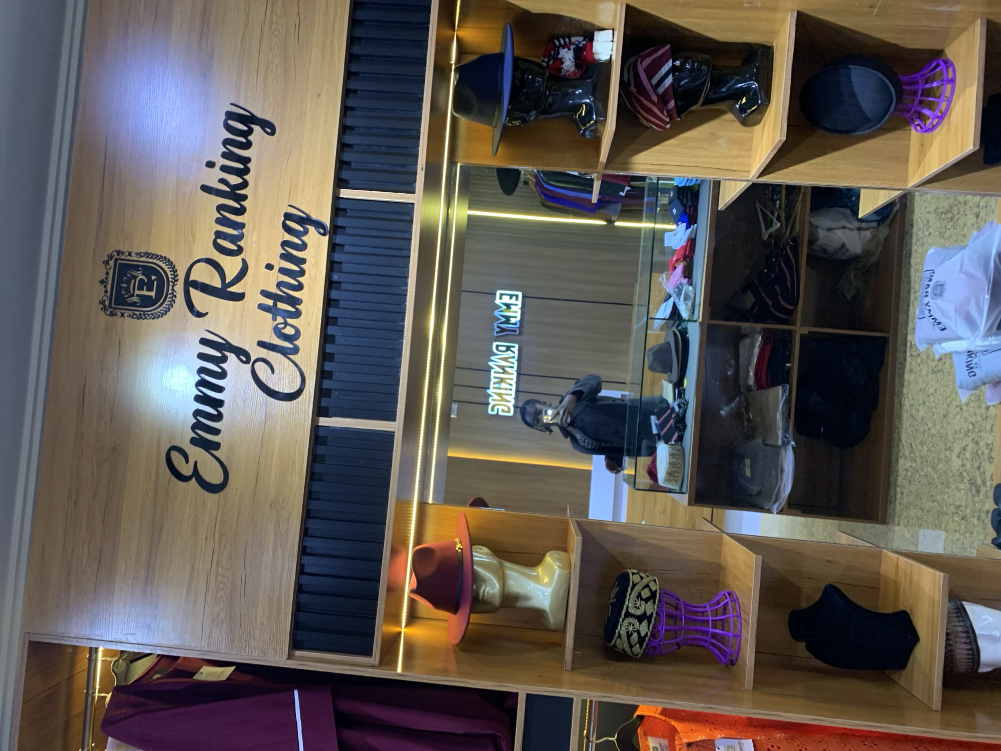 The image depicts the interior of a clothing store, possibly located in Gura Top, Nigeria, with shelves displaying various headwear and clothing items. The store features a prominent wooden sign with the words "Emmy Ranking Clothing" in black, stylized lettering, accompanied by a crest. On the left side of the store, a mannequin head wears a maroon fedora, and below it, another display showcases a black and gold embroidered cap on a purple stand. Further down, a black necklace display and a white mannequin head wearing a patterned cap are visible.
To the right, black mannequin heads are displayed with hats. One wears a black fedora with a blue band, and next to it is a display with a red and white striped cap. Below these, a black hat sits on a purple stand, and the bottom shelf shows a black cap.
In the background, a mirror reflects the store's interior. A person, likely a woman, is seen taking a selfie in the mirror, wearing a dark jacket. Behind her, clothing items hang on racks. A glass display case in the foreground contains several hats and other accessories. The overall environment appears to be a retail space with wooden shelving and good lighting, illuminated by what appears to be LED strips.