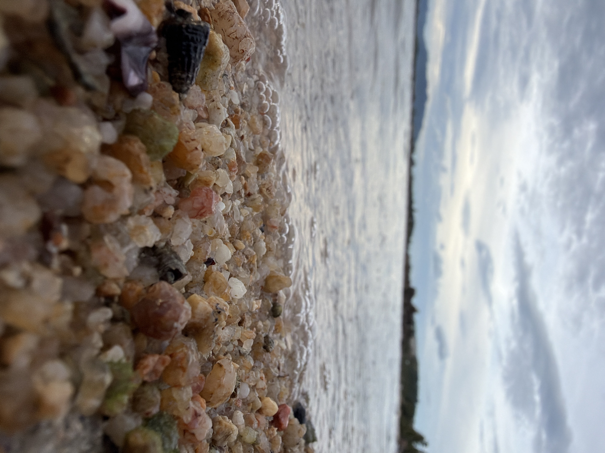 A low-angle, close-up shot captures a sandy beach in Koh Samui District, Thailand. The foreground is dominated by a bed of small, multi-colored pebbles and shells, with the edge of the water lapping gently at them, creating a line of small bubbles. Some larger, more distinct pebbles and shells are visible, including a dark, conical shell. Beyond the beach, the shallow water extends towards a blurred horizon where the muted tones of land and sky meet under an overcast, cloudy sky. The overall impression is one of a quiet, natural coastal scene.