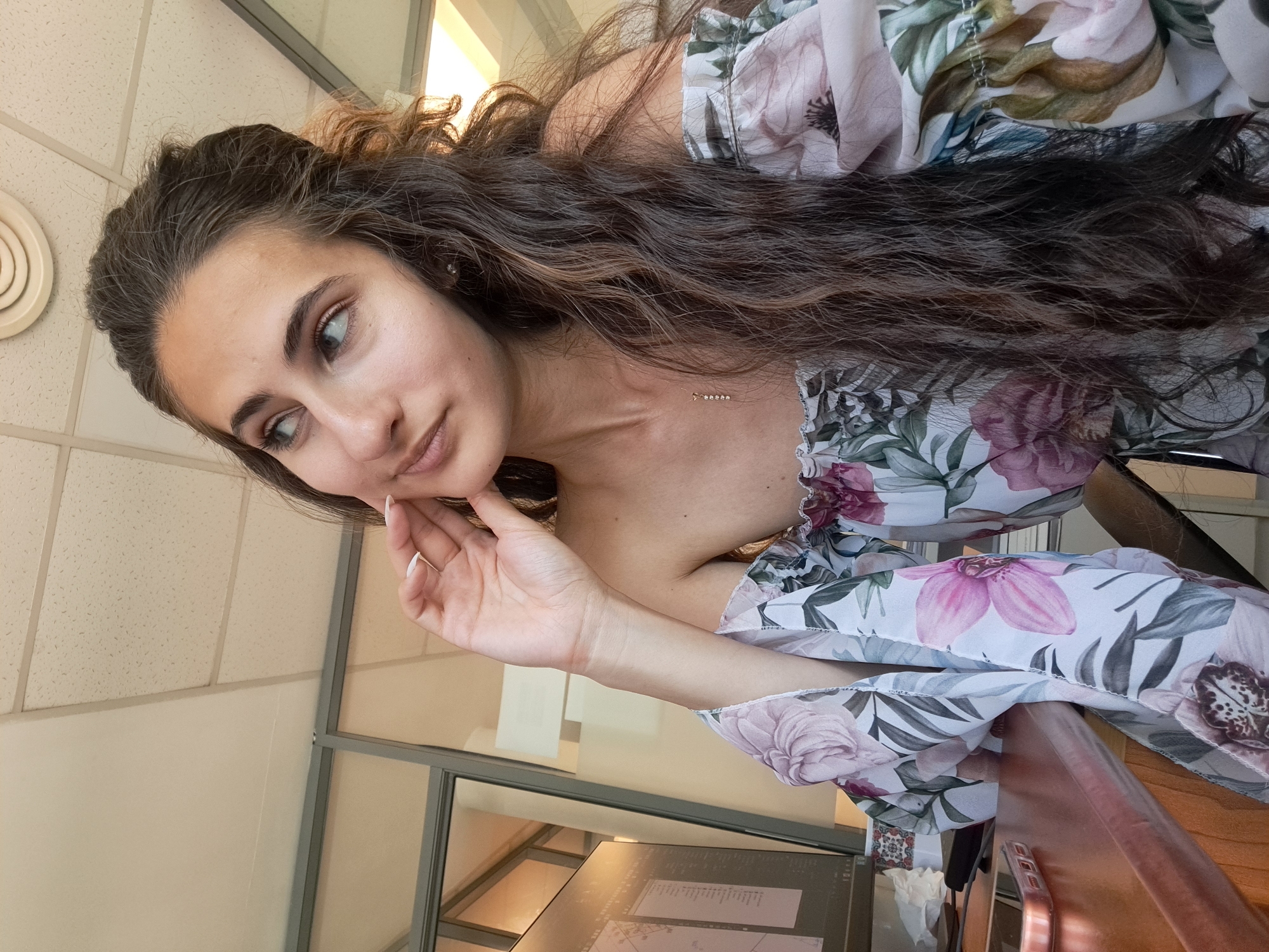 A young woman with long, dark, curly hair is seated at a wooden desk, facing towards the right side of the frame and looking away from the camera. She is wearing an off-the-shoulder floral print top with large pink and purple flowers. Her right hand is raised and touches her cheek. In the background, there is a computer monitor displaying data, a glass partition, and a ceiling with fluorescent lighting. The overall scene suggests an indoor office environment, possibly in Sofia, Bulgaria, given the provided context.