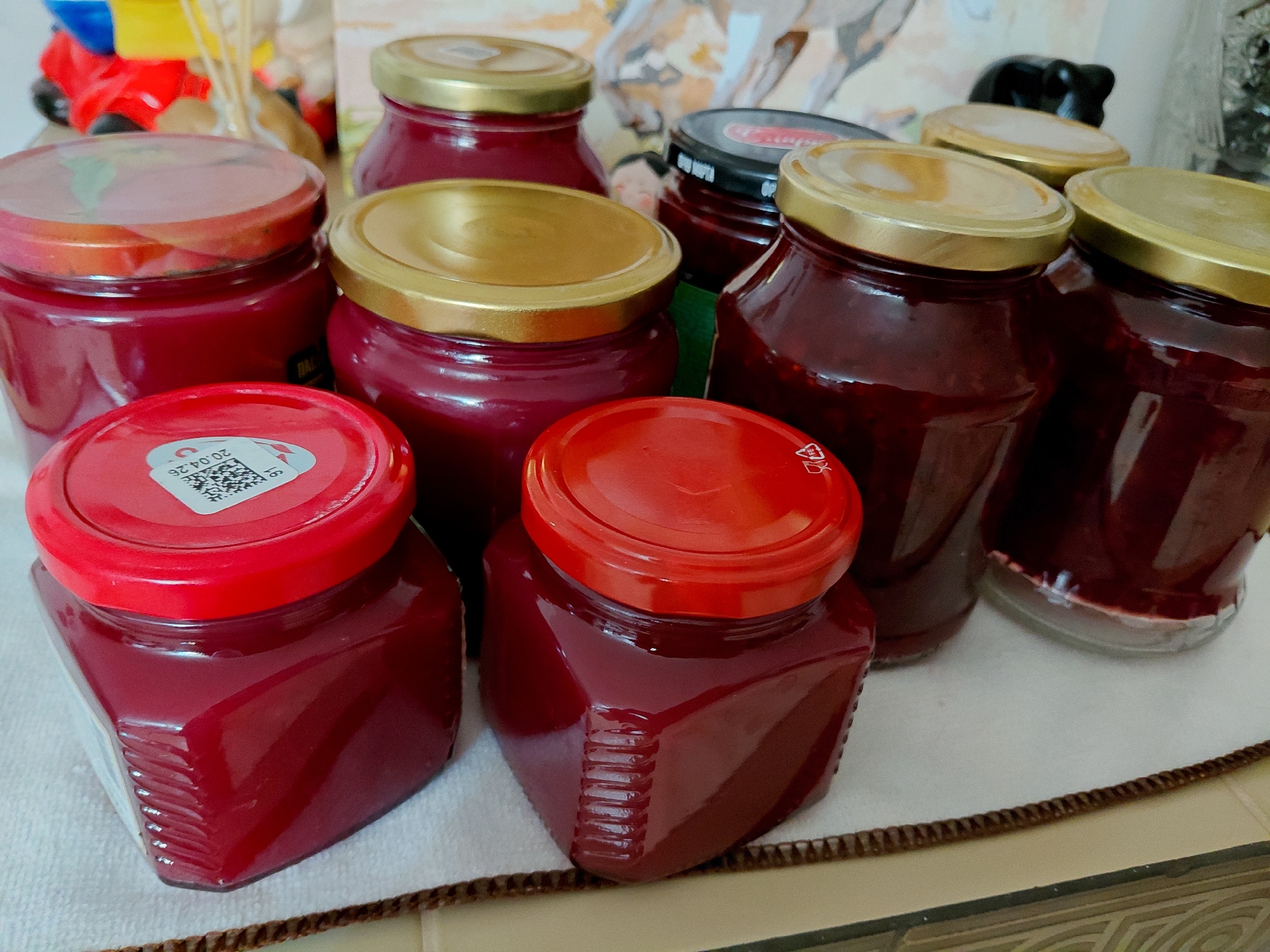 The image displays a collection of jars filled with a deep red, viscous substance, likely jam or preserves. The jars vary in size and shape, with several featuring metallic gold lids and two with red lids. One of the jars with a red lid has a white label with black text and a QR code, indicating a date of "20.04.26". The jars are arranged on a light-colored surface, possibly a tablecloth or mat, which is placed on a darker, textured surface. In the background, out of focus, are elements of what appears to be a home environment, including a blurry colorful object and a patterned wall hanging. There are no people or animals visible in the image. The scene is indoors, in what could be a kitchen or pantry. The specific location in the city of Сотниково, Russia, cannot be determined from the visual information provided.
