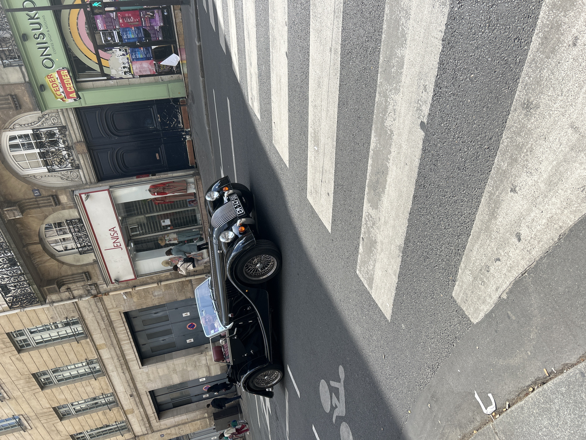 The image depicts a black vintage car parked on a crosswalk in an urban environment, likely in Paris, France. The car has the license plate "EM-147-DO". Pedestrians are visible on the sidewalk next to buildings with various shops and apartments. One shop is named "JENISA" and another named "ONISUKO COFFEE SHOP & MOCHIS" has posters on its storefront. The architecture of the buildings is characteristic of Parisian style.