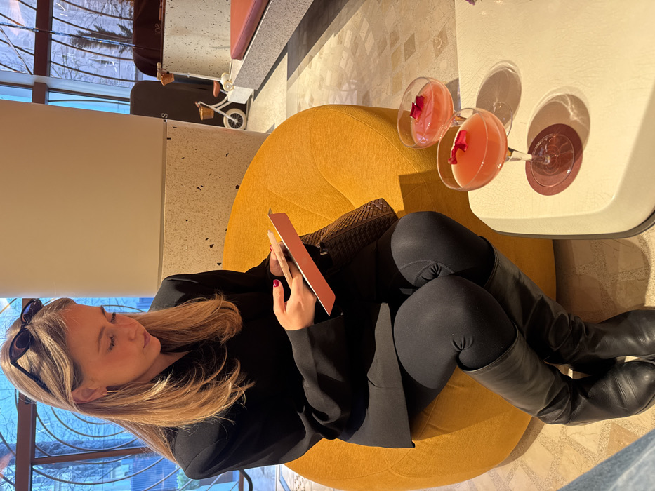A woman with light brown hair, wearing a black blazer, black leggings, and black knee-high boots, is seated on a large, round, mustard yellow lounge chair in a modern indoor setting. Sunglasses are positioned on her head. She is looking down, focused on a small, light-colored device held in her left hand, which rests on a small orange notebook. A dark brown woven bag is partially visible to her left on the chair. To her right, on a white rectangular table with rounded edges, are two coupette glasses filled with a pink liquid and garnished with red flower petals. One glass is placed on a round red coaster. The background includes large windows with a curved framework, showing blurred outdoor foliage. White structural columns, some with speckled bases, are also visible, along with a small white decorative bicycle near additional seating. The flooring is light-colored and reflective. The location is an establishment in Unknown, Unknown.