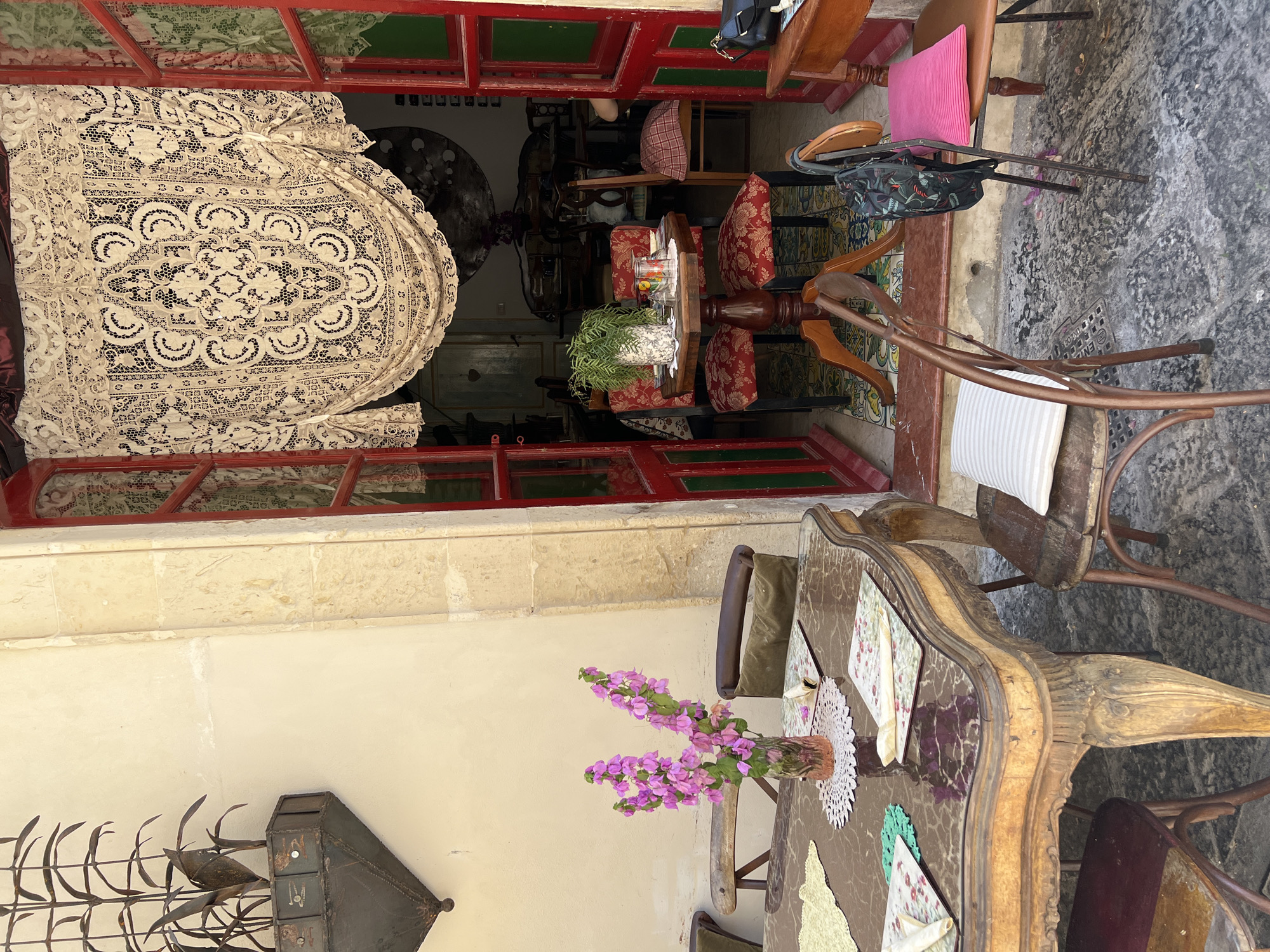 This image depicts an outdoor seating area of a cafe in Syracuse, Italy, likely located in the historic Ortigia district, known for its charming, narrow streets and ancient architecture. The scene is bathed in natural sunlight, highlighting the textures and colors of the vintage-inspired decor.
In the foreground, a large, ornate wooden table with a marble or dark polished top is set with placemats, napkins, and a vase of vibrant pink flowers. The table's legs are intricately carved, adding to its antique appeal. Several chairs surround the table, some made of dark wood with upholstered seats, and others of a more rustic metal construction. One chair, to the right of the table, has a bright pink cushion.
Behind the table, an open doorway leads into the interior of the cafe. The doorway is framed by bold red painted trim and adorned with a decorative lace curtain. Through the doorway, glimpses of tables and chairs are visible, suggesting a cozy indoor seating arrangement. The interior appears to be decorated with eclectic items, including what looks like a large circular artwork or mirror on the back wall.
To the left of the table, a portion of a cream-colored wall is visible, featuring a rustic, dark metal planter with stylized metallic foliage. The ground beneath the seating area is paved with a textured, uneven surface, possibly cobblestones or old flagstones, with scattered debris and a metal grate.
The overall atmosphere is one of rustic charm and bohemian elegance, inviting patrons to relax and enjoy a drink or meal in a unique setting. There are no people visible in the image, but the presence of table settings implies recent or anticipated customer activity.
