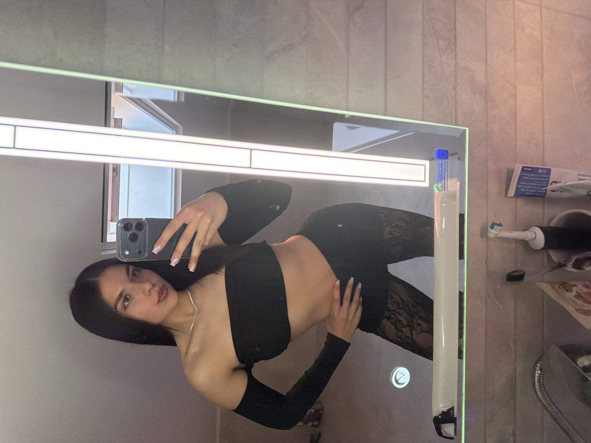 A young woman is taking a mirror selfie in what appears to be a bathroom. She is wearing a black crop top with long sleeves and black patterned pants. She is holding a smartphone in her left hand, with the screen facing the mirror. Her nails are manicured with a white design. The background includes a mirror with an integrated light, a tiled wall, a toothbrush, toothpaste, and other bathroom items. The location in Gabrovo, Bulgaria, is not discernible from the image.