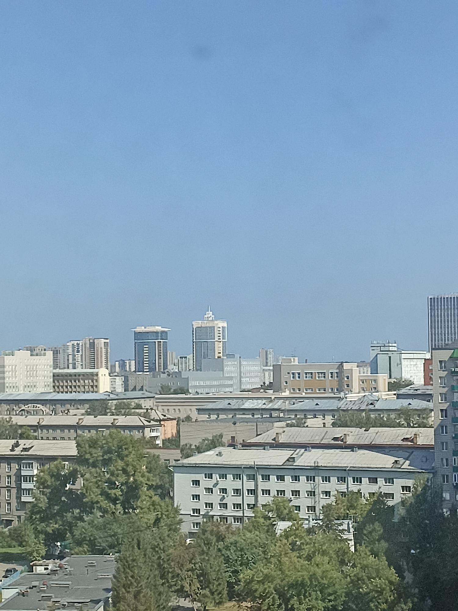 The image depicts a cityscape of Novosibirsk, Russia, under a clear blue sky. The foreground features lush green trees partially obscuring several apartment buildings with numerous windows, typical of Soviet-era architecture. In the mid-ground, a series of low-rise buildings with flat roofs are visible, some showing signs of wear. Beyond these, a more modern urban landscape emerges with taller buildings, including high-rise apartment complexes and office buildings, some with distinctive architectural features like rounded corners or glass facades. The tallest structures in the distance create a skyline that suggests a significant urban center. The overall impression is that of a densely populated city with a mix of older and newer construction.