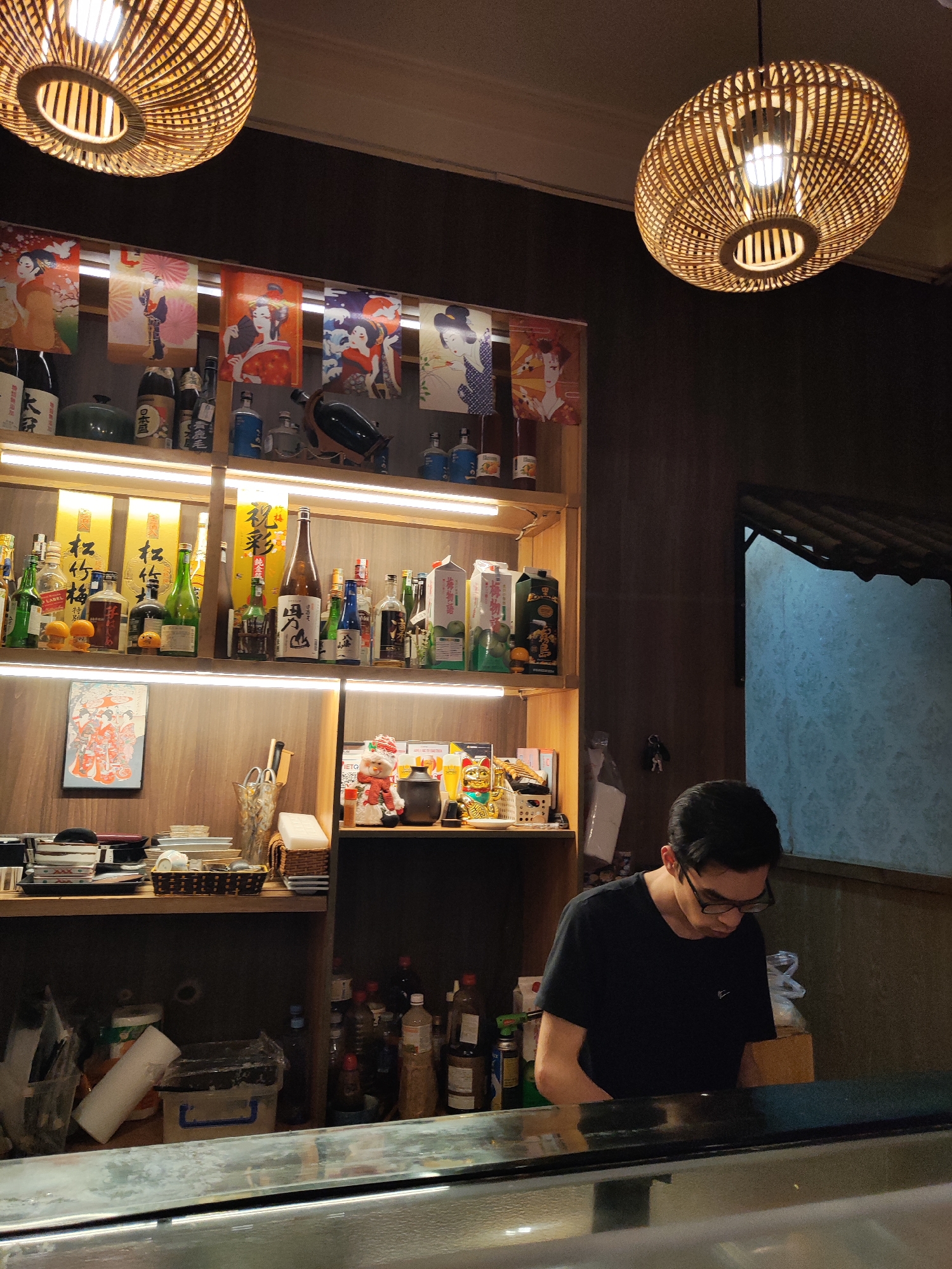 A person is working behind a counter in an establishment that appears to be a sushi restaurant, as suggested by the general decor and items displayed on the shelves behind them. The shelves are stocked with various bottles, likely including sake, and Japanese-themed decorations, such as posters featuring geishas and other traditional imagery. The counter is in the foreground, partially obscuring the view of the worker. The location is unknown. Two wicker light fixtures hang from the ceiling, providing illumination to the scene. A window is visible on the right side of the image.