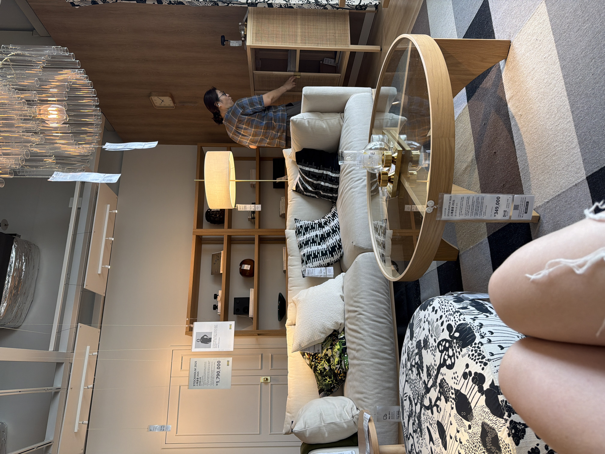 This image depicts an interior scene at an IKEA store in Goyang-si, South Korea. In the foreground, a portion of a person's legs is visible, seated on a circular, patterned ottoman. The ottoman features a black and white floral design. In the mid-ground, a light-colored sofa with several decorative pillows is arranged, including one with a green and black abstract pattern and another with a black and white geometric design. A round glass-topped coffee table with a light wood base sits in front of the sofa, holding a tray with two gold candle holders and a glass beaker. The background reveals a wooden shelving unit filled with various items, including books and decorative objects. A floor lamp with a light-colored shade stands to the left of the shelving unit. A woman in a plaid shirt is visible in the right mid-ground, opening a cabinet with woven rattan detailing. A clock is mounted on the wood-paneled wall behind her. The ceiling features a modern chandelier made of glass tubes. Several IKEA price tags and informational posters are hanging from the ceiling and attached to furniture. The floor is covered with a patterned rug in shades of gray, beige, and brown.