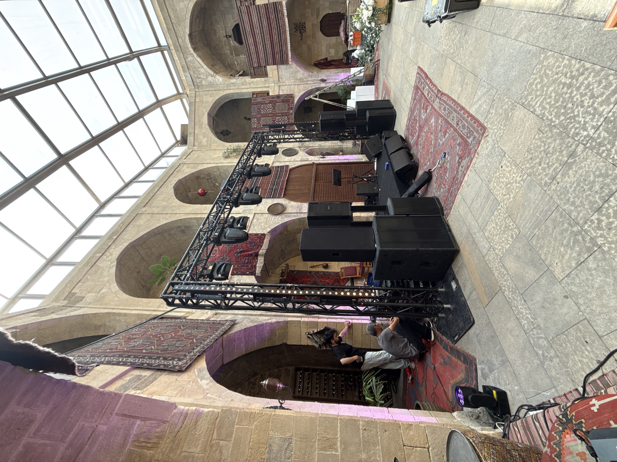 In the Sabail Raion, Azerbaijan, a courtyard with stone flooring and arched architecture features a stage setup. The stage consists of a black platform, a truss structure holding multiple lights, and several large black speakers. Two men are present, one in a black shirt and grey pants, and another in a grey t-shirt and dark pants, appearing to be working on the stage equipment. Decorative Persian rugs are hung on the stone walls and spread on the floor. The courtyard is covered by a glass ceiling, allowing natural light to enter.
