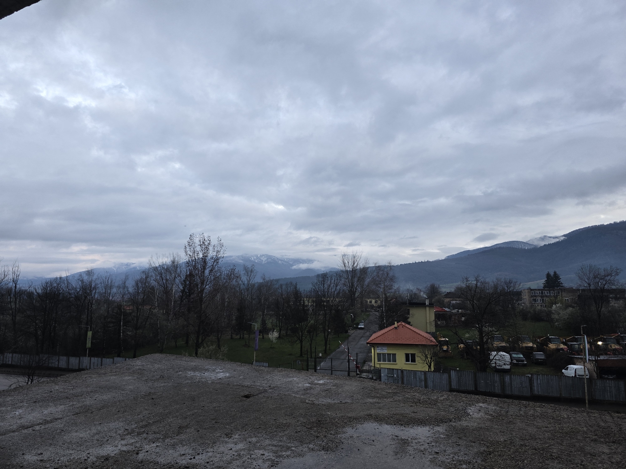 The image depicts a cloudy scene in Berkovitsa, Bulgaria. The foreground features a rough, gravelly surface, possibly a construction site or undeveloped area. Beyond this, a view opens up to a landscape with trees, a yellow building with a red tiled roof, and a fenced area containing various vehicles, including vans and what appear to be construction vehicles. In the background, a mountain range is visible, partially covered in clouds and possibly with some snow on the peaks. The sky is overcast with a thick layer of gray clouds. The overall atmosphere suggests a cool or cold, possibly damp day.