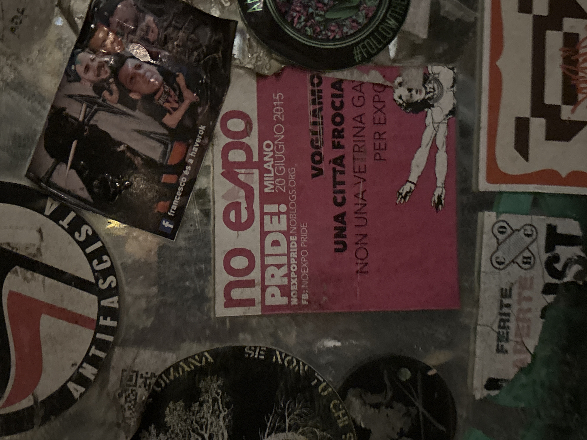 This is a close-up image of a collection of stickers adhered to a surface. The stickers include:
* A sticker with a group of people, identified as "francesco és a haverok" with a Facebook logo.
* A sticker with "no expo PRIDE!" and "Milano 20 Giugno 2015". The text also includes "NOEXPOPRIDE.NOBLOGS.ORG" and "FB: NOEXPO PRIDE".
* A pink sticker with the text "VOGLIAMO UNA CITTÀ FROCIA NON UNA VETRINA GA PER EXPO" accompanied by a drawing of a person.
* A sticker with "ANTIFASCISTA" arranged in a circular pattern.
* A sticker saying "SE NON TU CHI S...".
* A sticker with "\#FOLLOWTH".
* A sticker with "FERITE APERTE ST".
The stickers appear to be attached to a metal surface. The specific location in Milan is not visually discernible from the stickers.