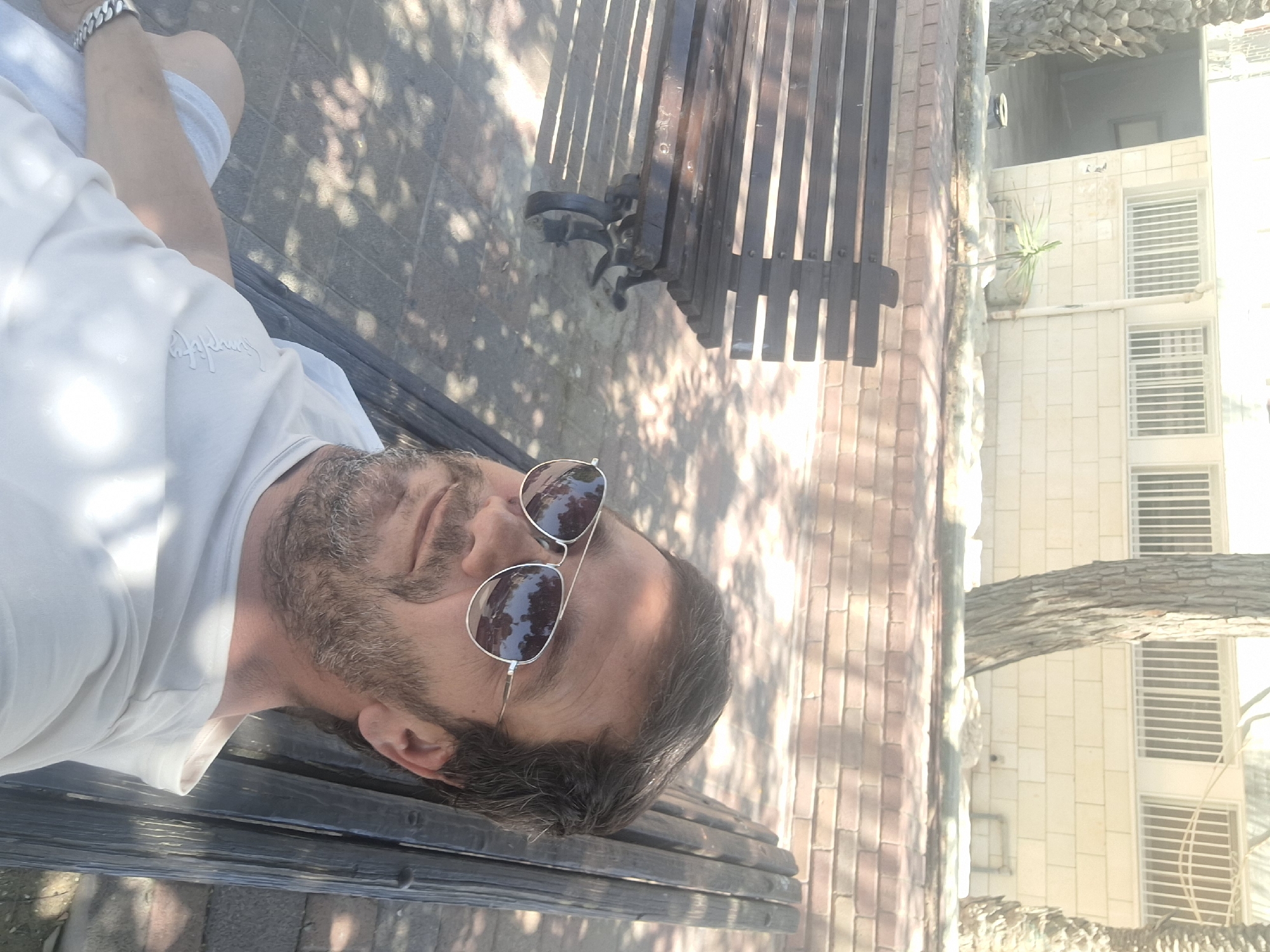 A man with short brown hair and a beard wearing a white t-shirt and sunglasses is sitting on a dark wooden bench. He is in a public outdoor space with a brick wall and a building in the background. The area appears to be Yerocham, Israel, given the architectural style of the building and the presence of palm trees. The sun is shining, casting shadows on the ground. The man is looking at the camera, and his reflection is visible in his sunglasses, showing trees and buildings.