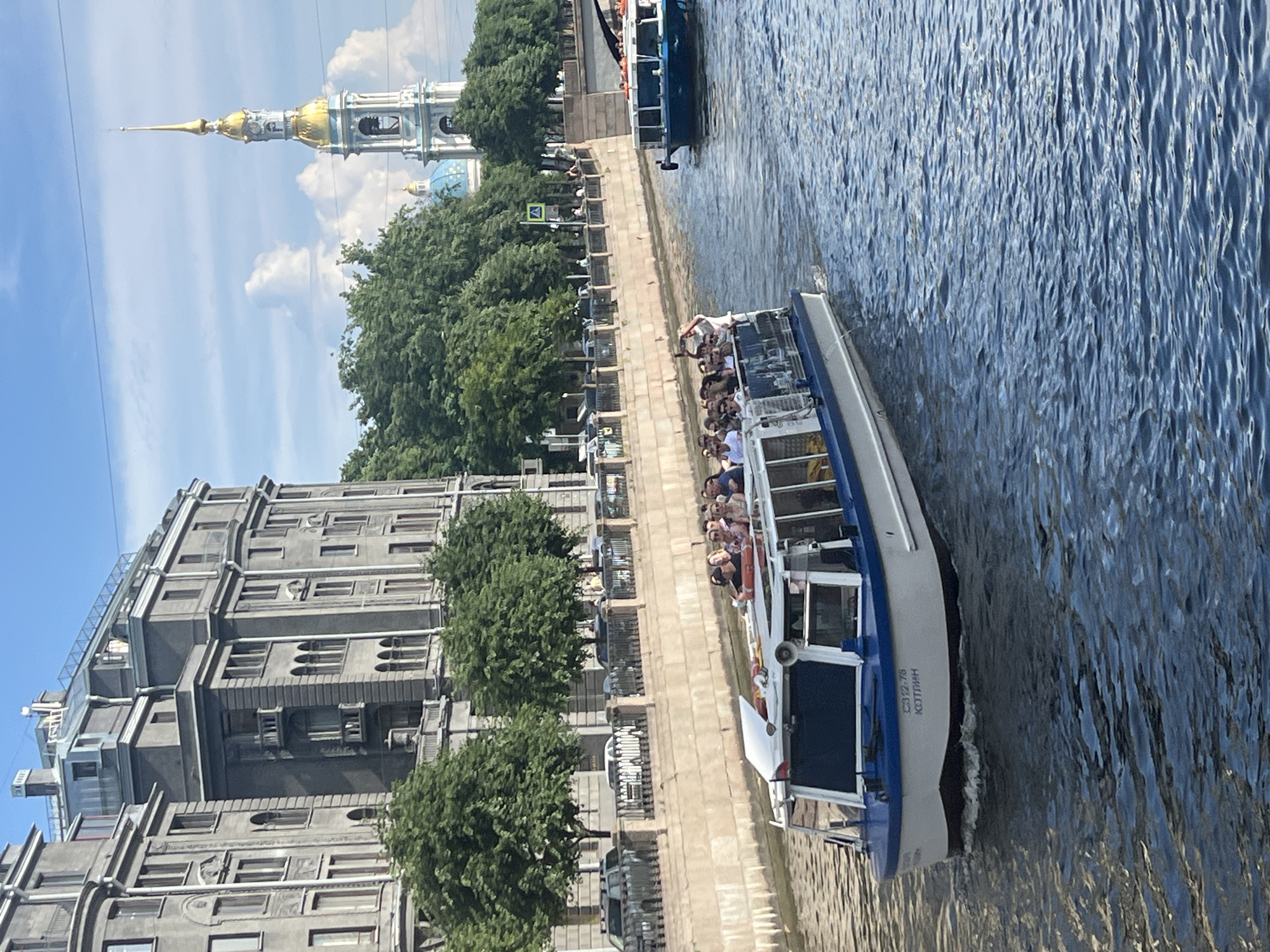 A wide shot captures a canal in Saint Petersburg, Russia, on a bright, partly cloudy day. A white and blue tour boat, filled with passengers, navigates the water in the foreground. The boat bears the inscription "С312-78 КОТЛИН" on its hull. On the left bank, an imposing, multi-story building with ornate architecture lines the canal, flanked by mature trees. A black metal fence runs along the embankment. In the background, the iconic golden spire of a church, likely St. Nicholas Naval Cathedral, rises above the trees, its blue dome also visible. Another, smaller boat is visible further down the canal on the right. The water is a deep blue, reflecting the sky and the surrounding structures. The scene depicts a leisurely summer day in the city, with tourists enjoying a boat tour.